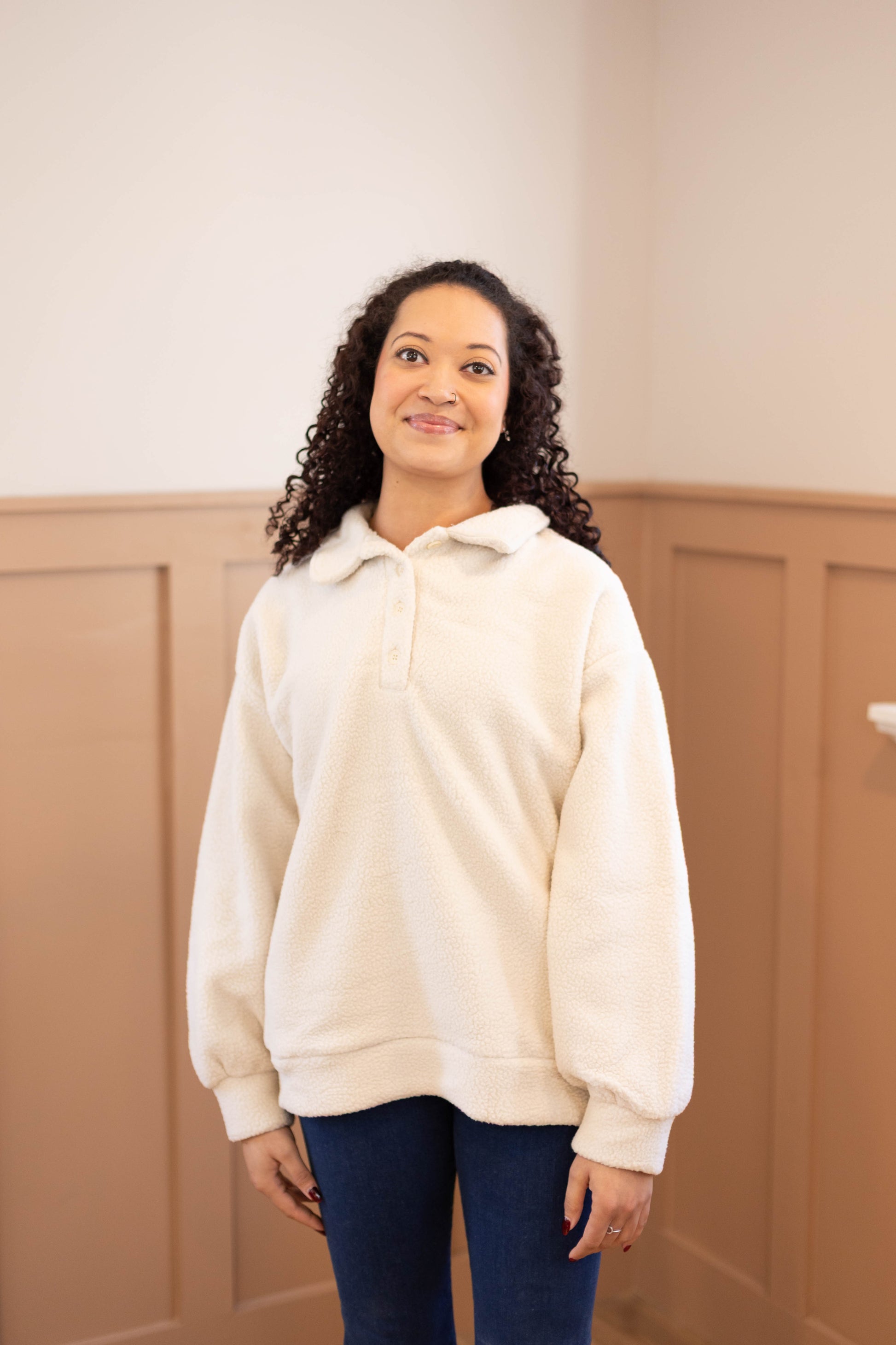 A woman with curly hair stands indoors, wearing the Dogwood Cloth Alex Sherpa Henley and blue jeans, in front of neutral-colored paneled walls.