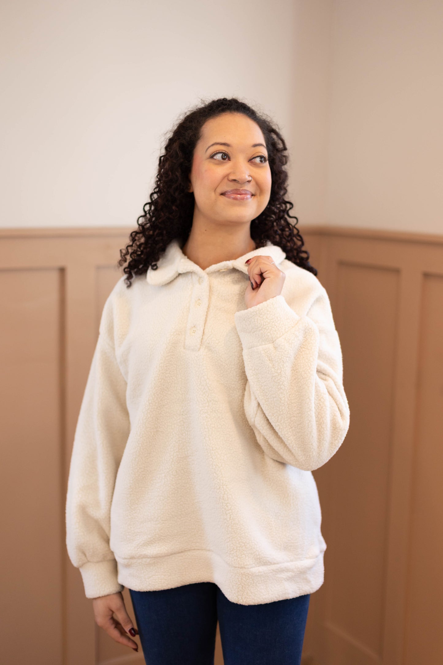 A woman with curly hair smiles indoors, wearing the Dogwood Cloth Alex Sherpa Henley in a relaxed fit with blue jeans, looking slightly to the side.
