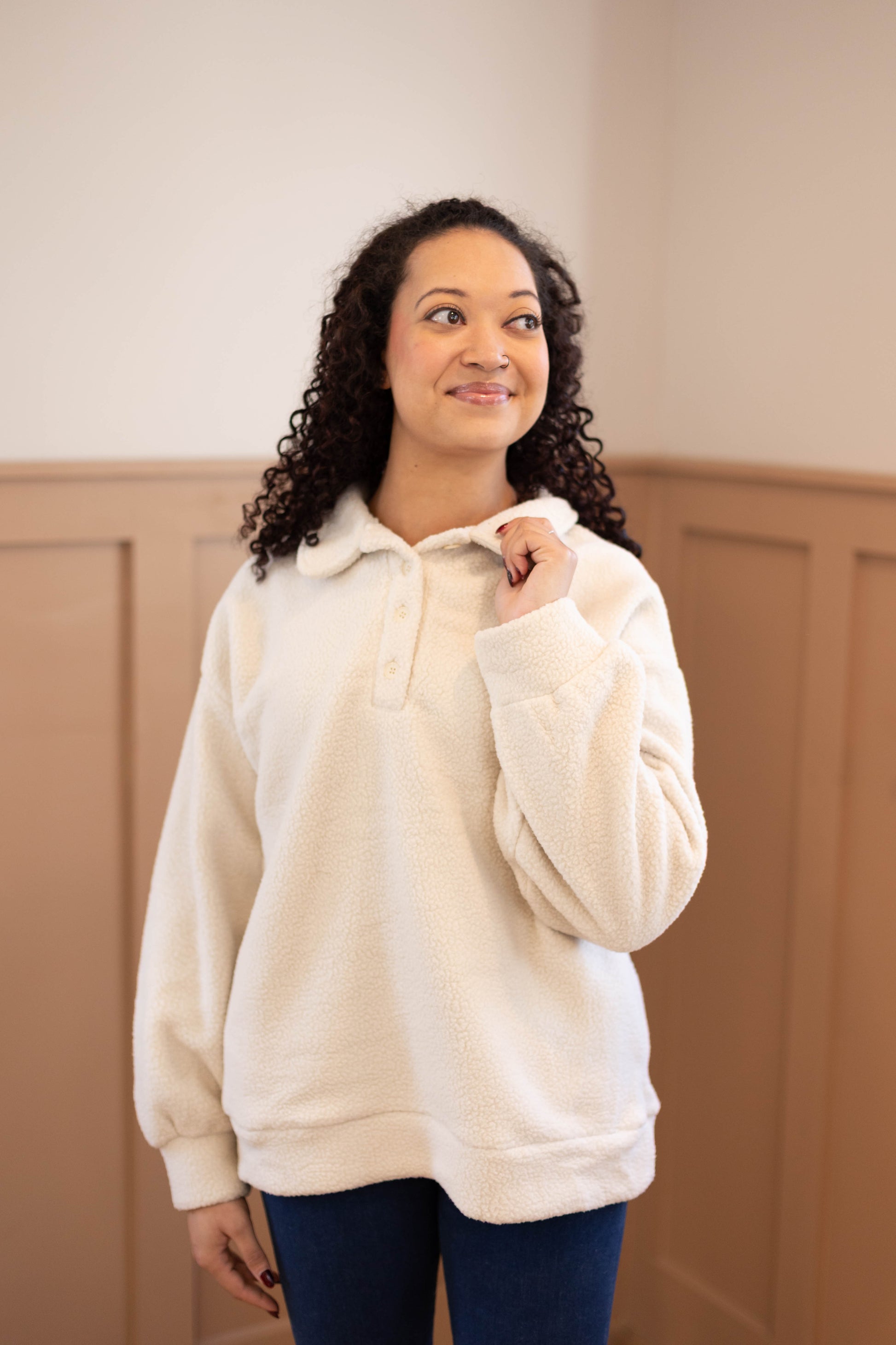 A woman with curly hair smiles indoors, wearing the Dogwood Cloth Alex Sherpa Henley in a relaxed fit with blue jeans, looking slightly to the side.