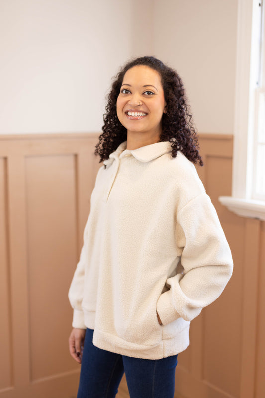 A woman with curly hair stands indoors, smiling at the camera. She wears the Dogwood Cloth Alex Sherpa Henley with jeans, her hands in her pockets.