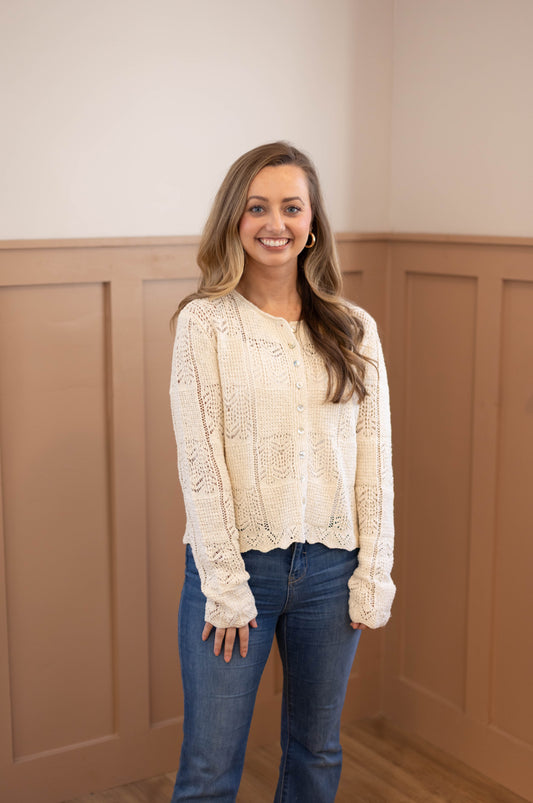 A woman with long hair smiles indoors, wearing the Dogwood Cloth Claire Lace Sweater in cream and blue jeans, framed by wooden paneling.