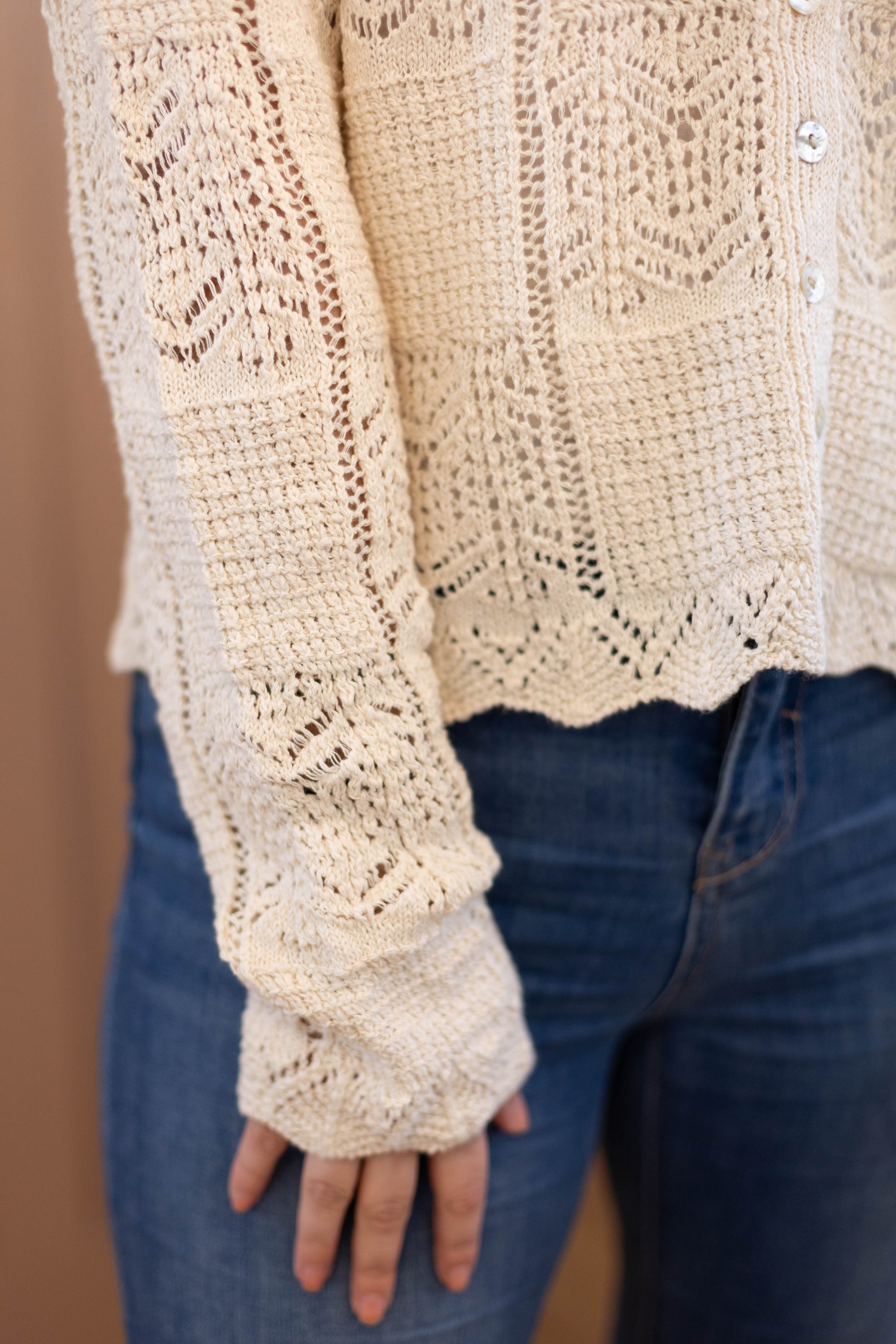 Close-up of a person wearing the Claire Lace Sweater by Dogwood Cloth, a cream cotton-polyester top with a scalloped hem, paired with blue jeans. Only the lower torso, right arm, and hand are visible. Hand wash cold; cool iron if needed.