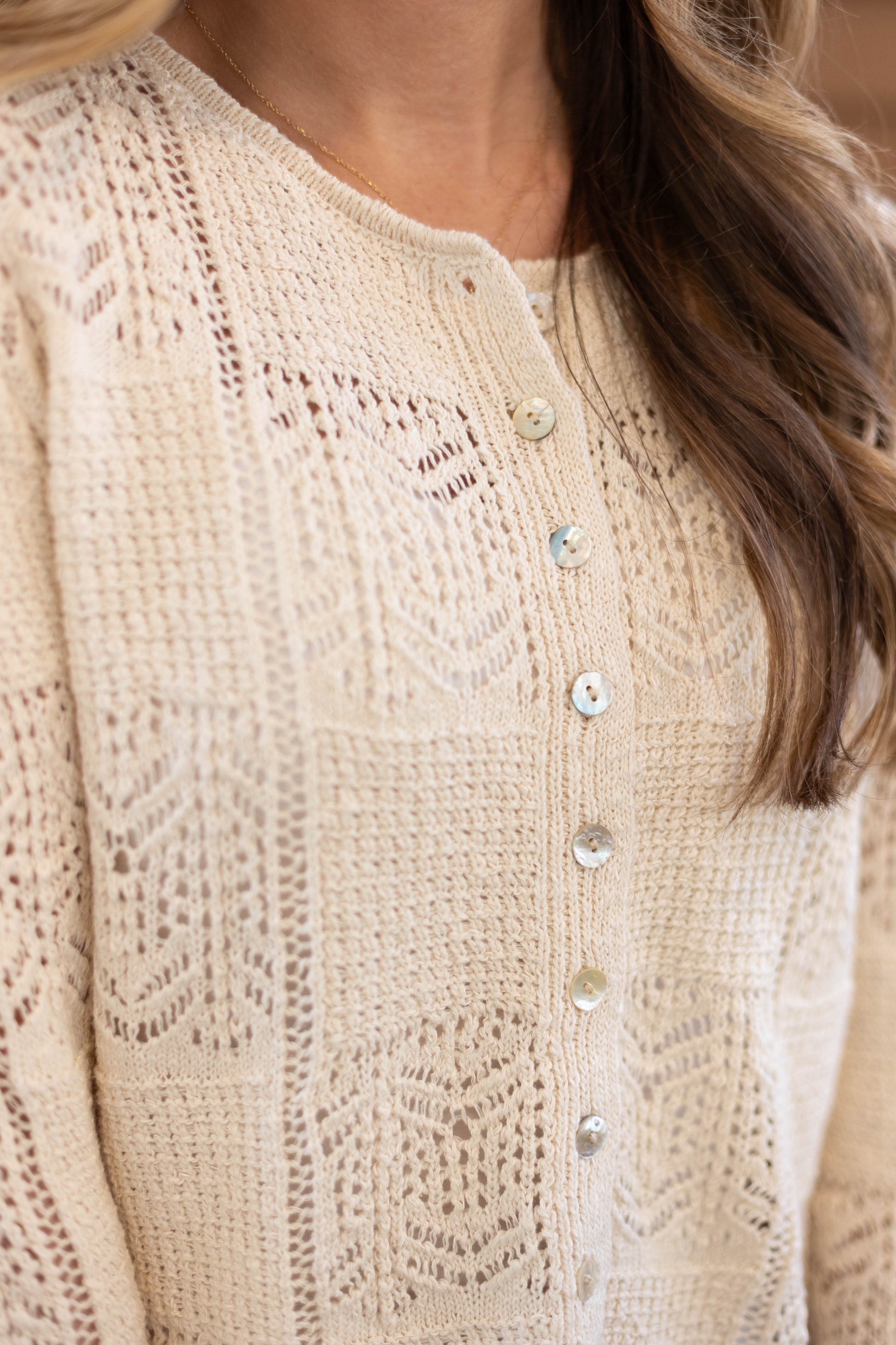 Close-up of a person wearing the cream Claire Lace Sweater from Dogwood Cloth, featuring a textured knit pattern and shiny buttons. For best care, hand wash cold and use a cool iron.