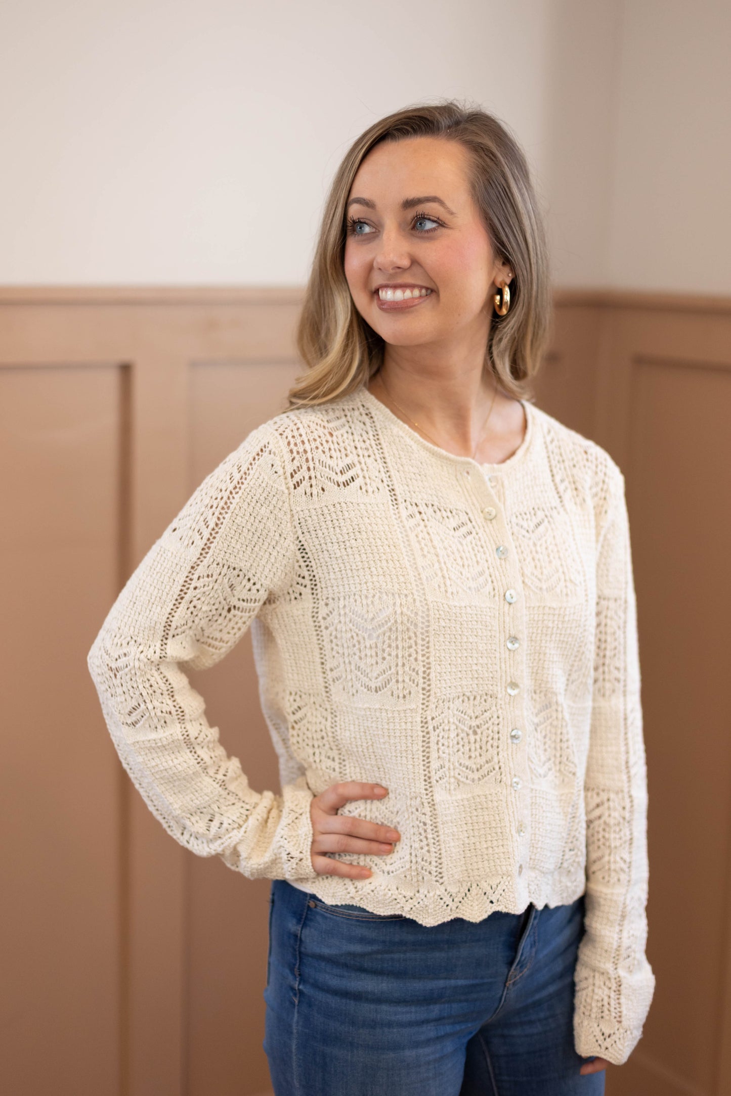 A woman wears the Claire Lace Sweater by Dogwood Cloth—cream, button-up, cotton-polyester blend with a women's fit—paired with blue jeans indoors, smiling to the side. Hand wash cold for best care.