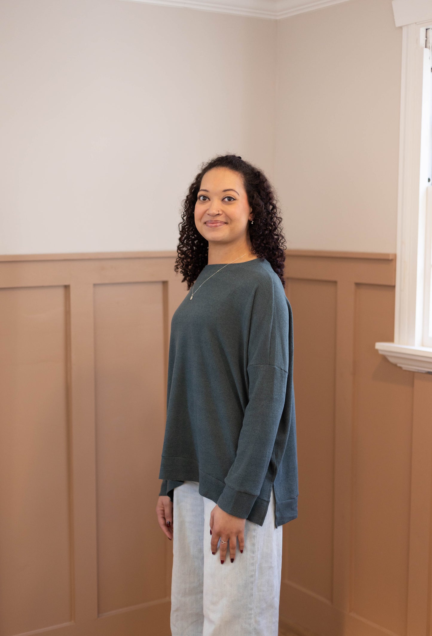 A woman with curly hair wears the Dogwood Cloth Chandler Sage Top and light jeans, standing indoors by a window and facing slightly to the side.