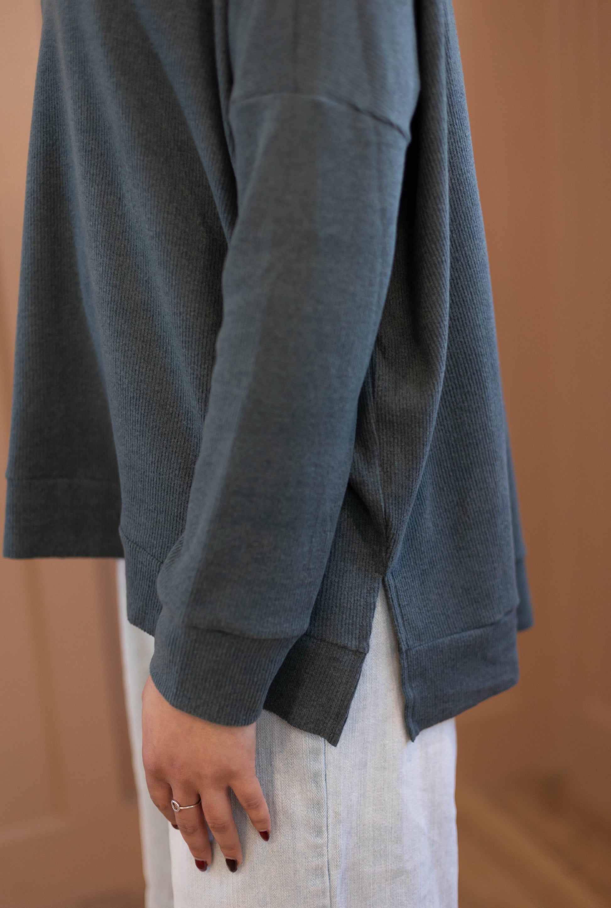 A person wearing the Chandler Sage Top by Dogwood Cloth, a cozy blue women’s sweater with a side slit, paired with light-colored pants and standing indoors with their arm relaxed at their side.