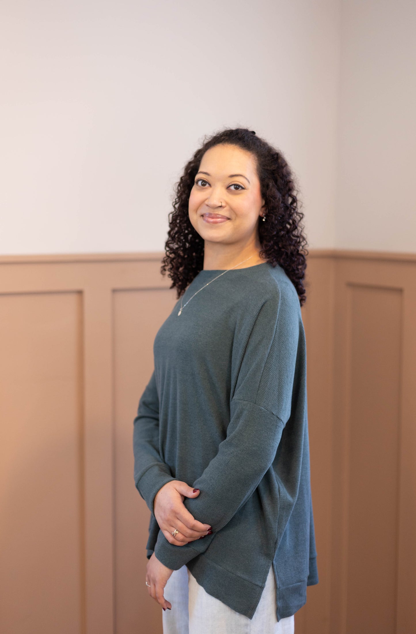 A woman with curly hair wears the Dogwood Cloth Chandler Sage Top indoors, smiling softly at the camera, with tan wainscoting in the background.