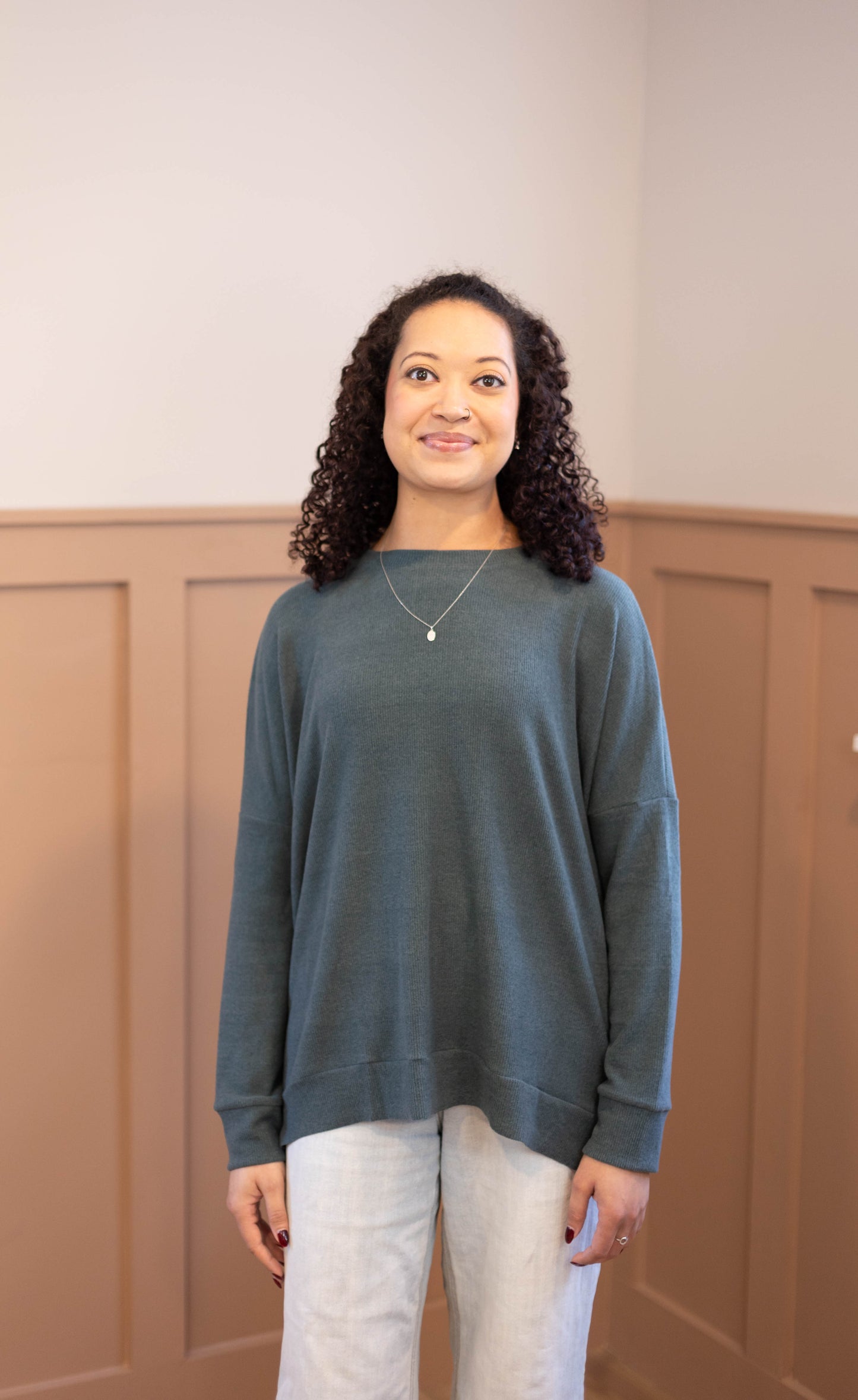 A woman with curly hair stands indoors against tan paneled walls, wearing the Dogwood Cloth Chandler Sage Top and light pants.