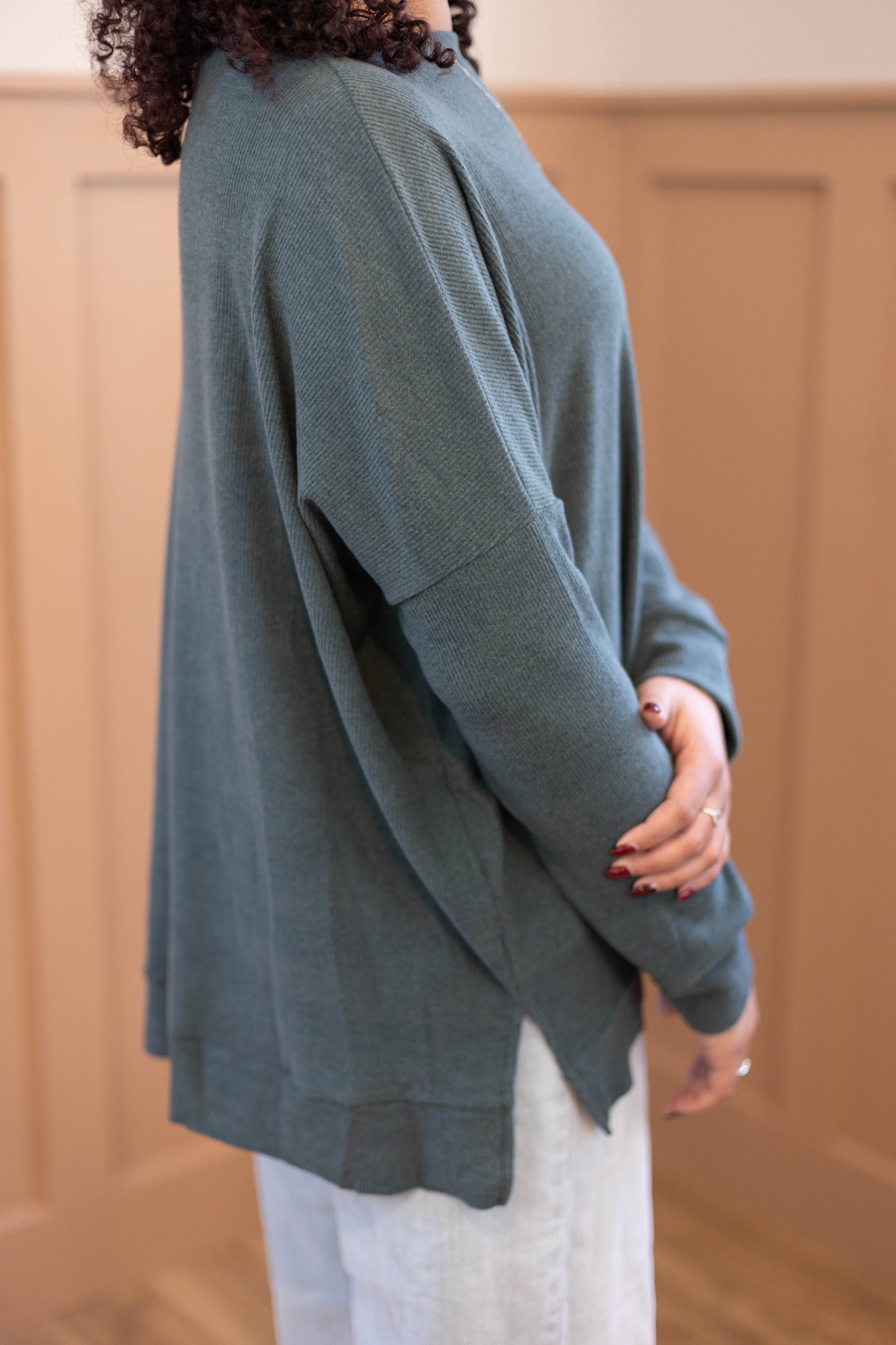 Someone stands sideways against a tan paneled wall, wearing a loose-fitting Chandler Sage Top from Dogwood Cloth with light-colored pants.