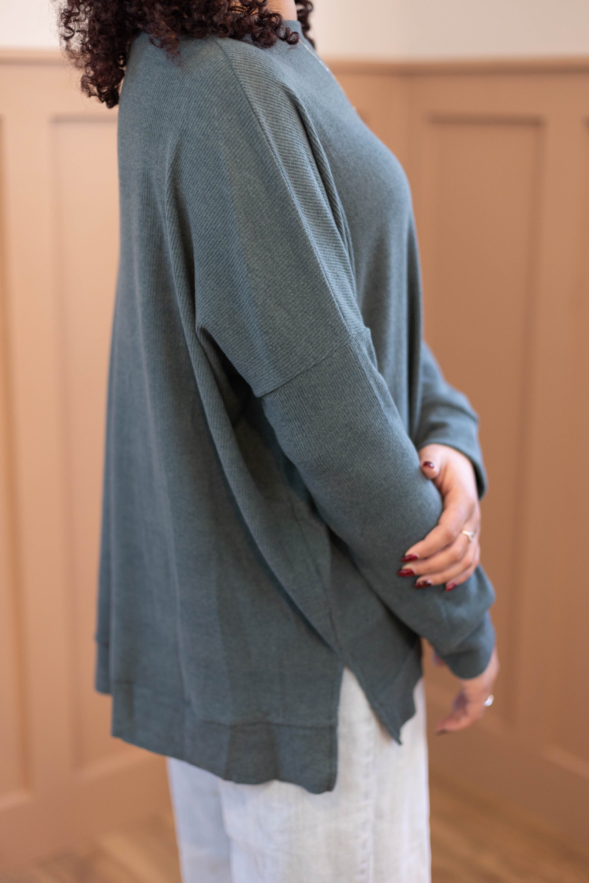 Someone stands sideways against a tan paneled wall, wearing a loose-fitting Chandler Sage Top from Dogwood Cloth with light-colored pants.