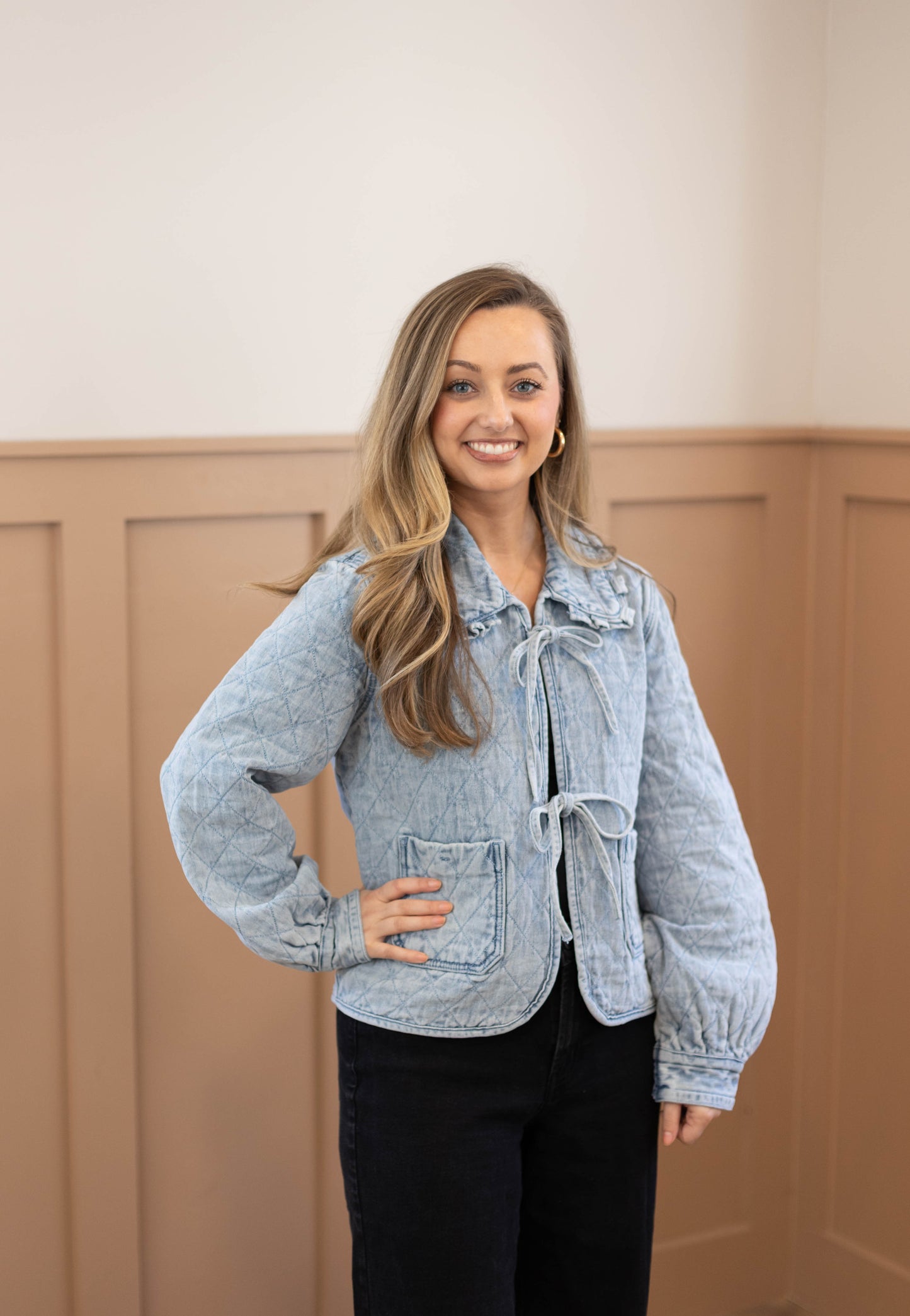 A woman with long blonde hair smiles at the camera while wearing the Dogwood Cloth Emma Quilted Jean Jacket and black pants, standing indoors against a beige paneled wall.