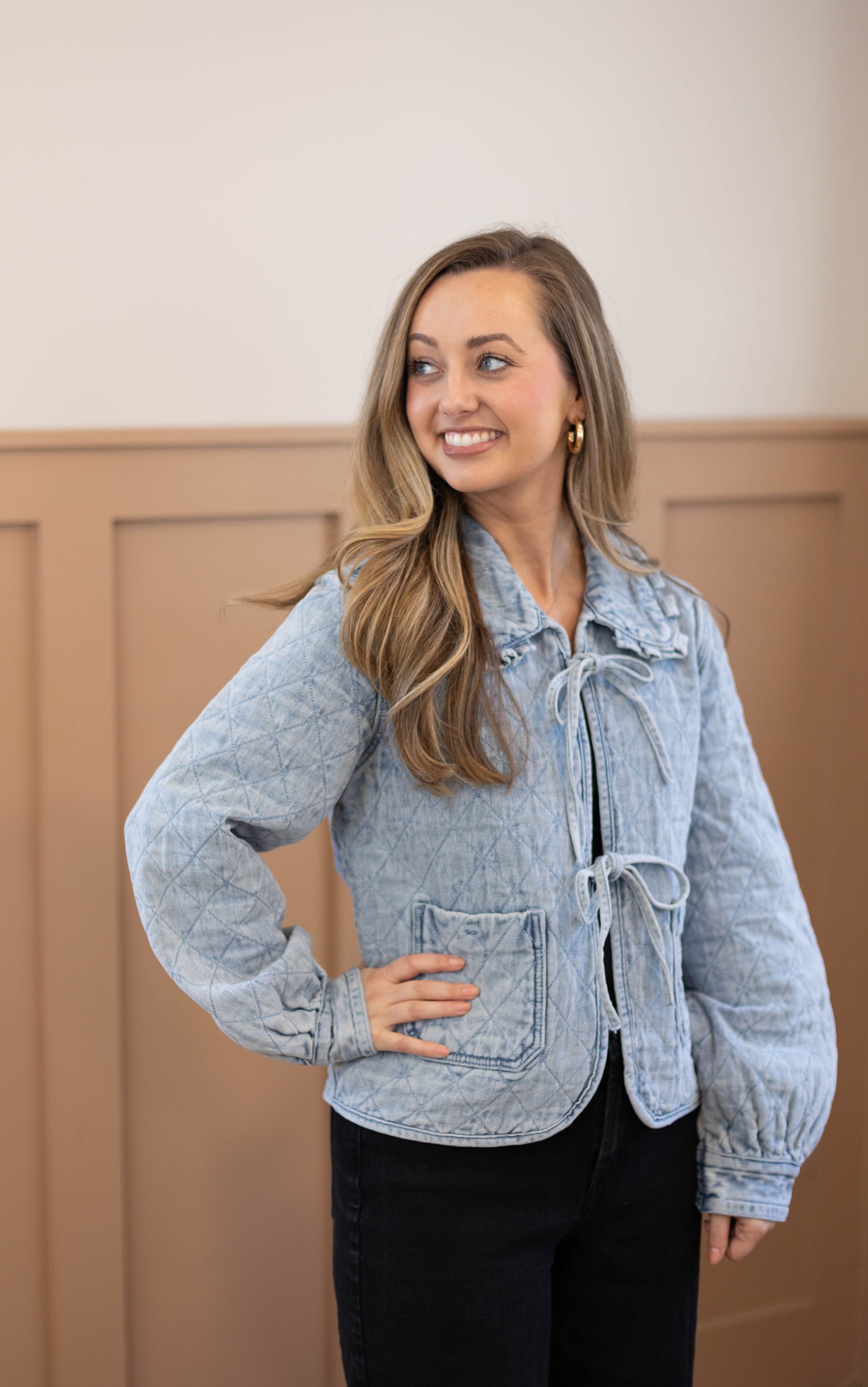 A woman stands indoors smiling and looking to the side, wearing the Dogwood Cloth Emma Quilted Jean Jacket in light blue with black pants.