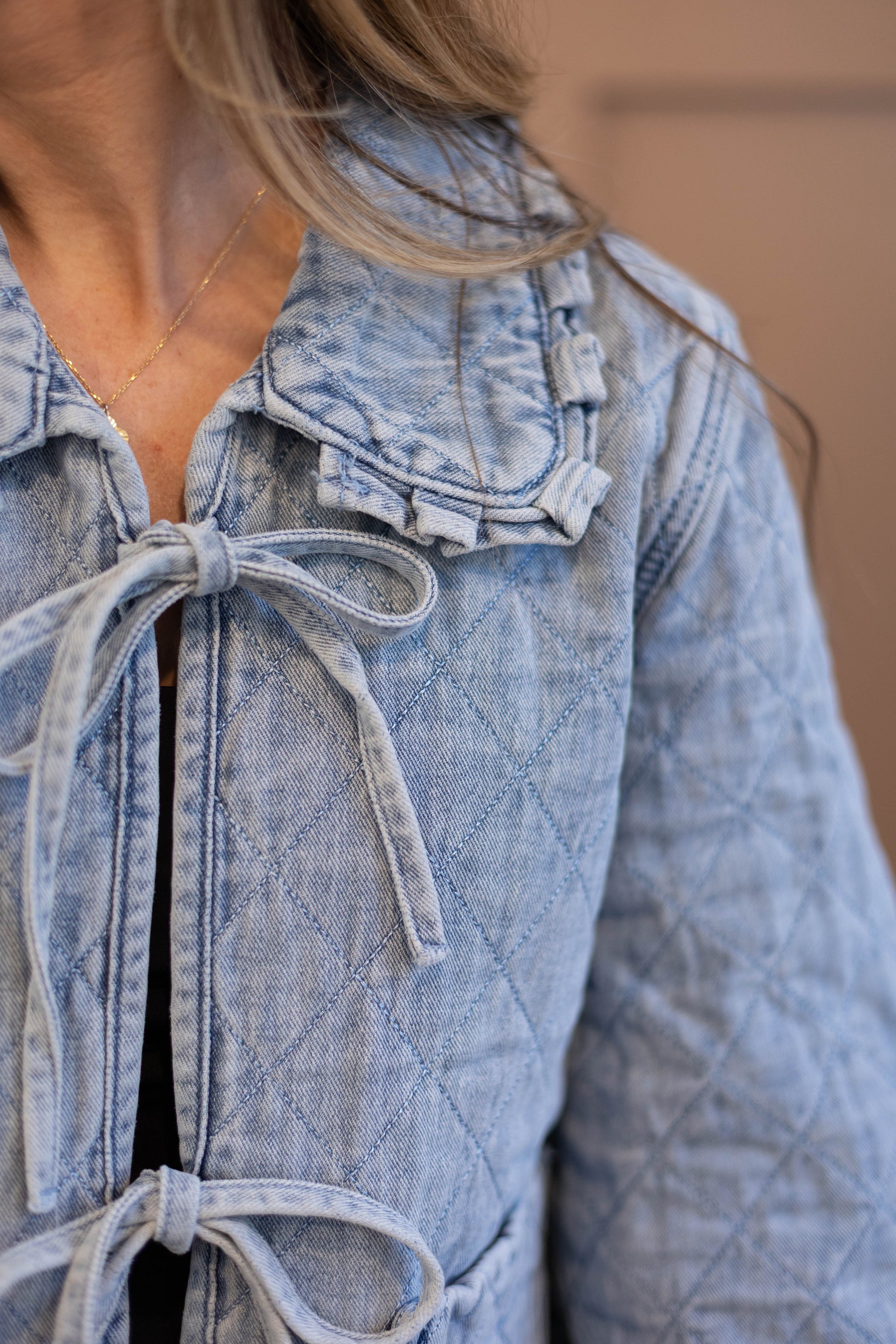 Close-up of a person wearing the Emma Quilted Jean Jacket by Dogwood Cloth, featuring light blue denim, quilted texture, and tie closures, paired with a gold necklace.
