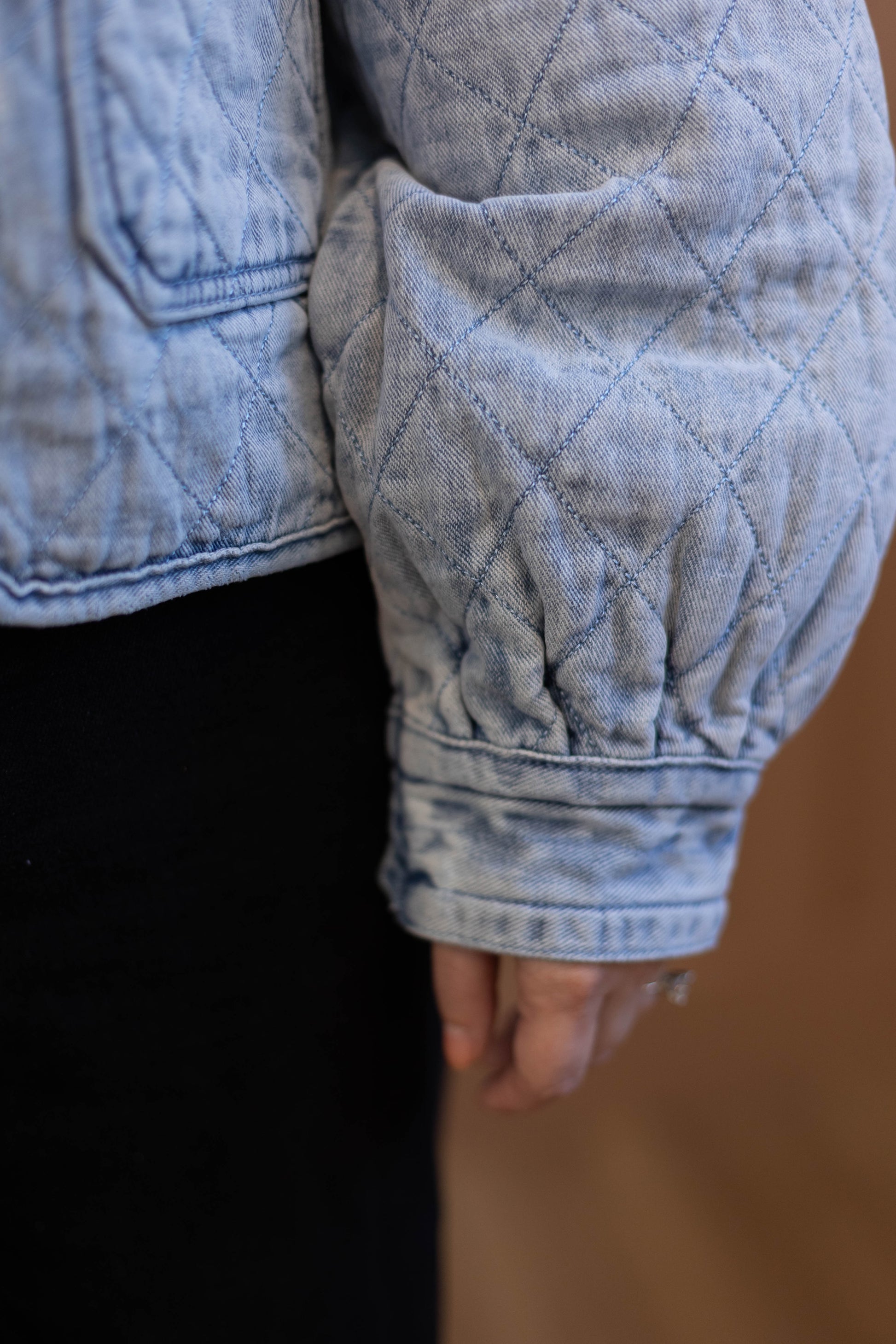 Close-up of a person wearing Dogwood Cloth's Emma Quilted Jean Jacket in light blue with gathered sleeves and black pants, showing part of their hand. The jacket’s soft texture adds a stylish touch to the look.