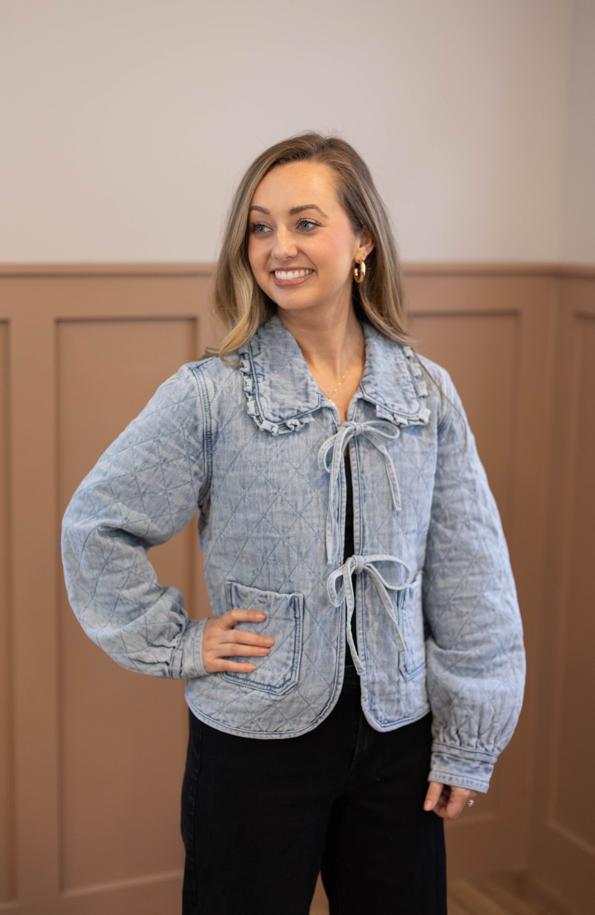 A woman indoors wears the Emma Quilted Jean Jacket by Dogwood Cloth, featuring a light blue hue, Peter Pan collar, and front ties. She pairs it with black pants, smiling as she looks to the side.