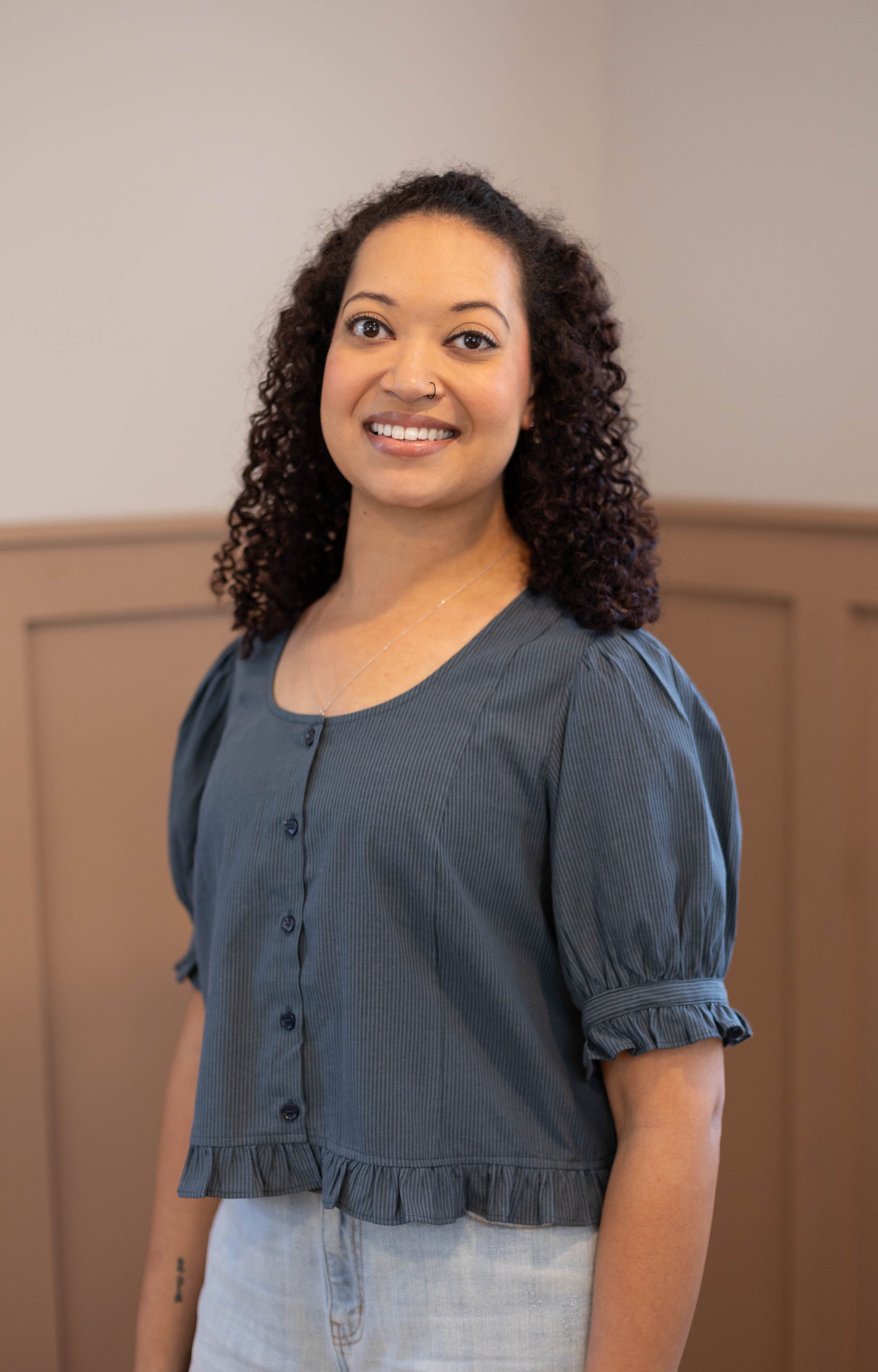 A woman with curly hair wears the Dogwood Cloth Millie Pinstripe Top in blue and light jeans, standing indoors before a beige paneled wall and smiling at the camera.