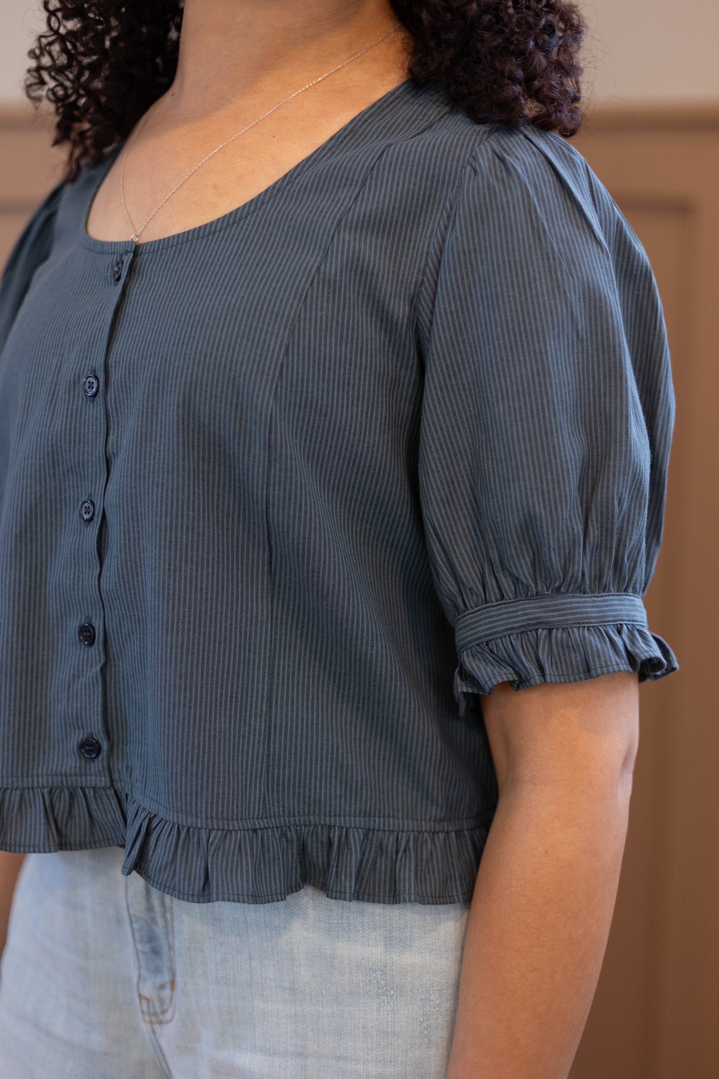 A person wears the Dogwood Cloth Millie Pinstripe Top, a blue short-sleeved feminine blouse with a ruffled hem, paired with light wash jeans, shown from shoulders to waist.