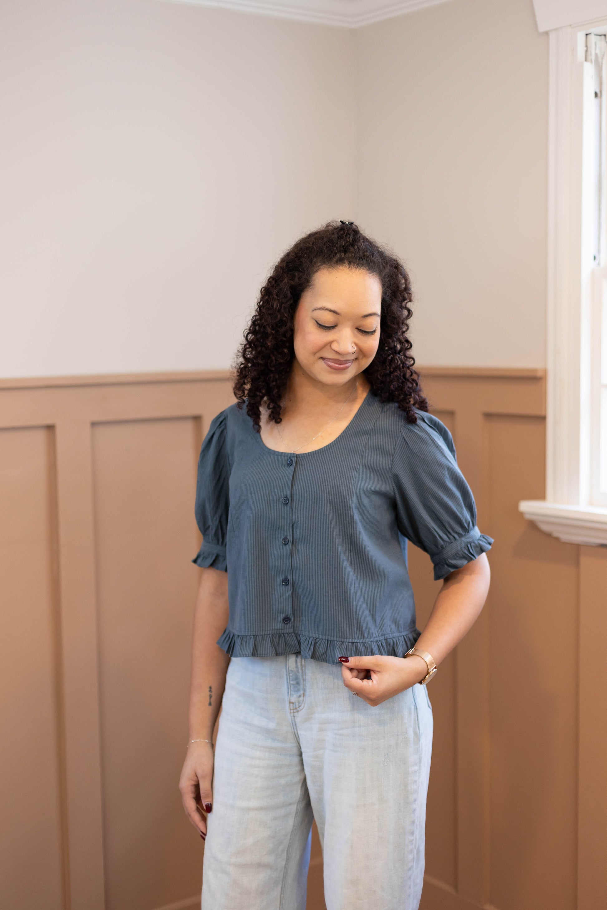 A woman with curly hair wears the Dogwood Cloth Millie Pinstripe Top, a feminine blue short-sleeve blouse, paired with light jeans as she stands indoors by a window with tan paneled walls.
