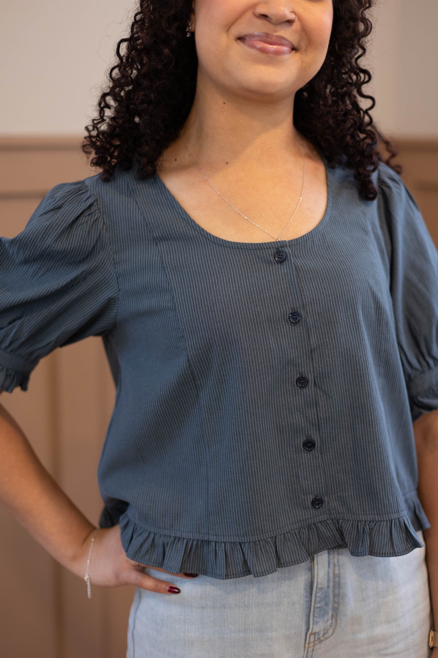 A person wears the Dogwood Cloth Millie Pinstripe Top, a blue short-sleeved feminine blouse with a ruffled hem, paired with light-wash jeans and standing with one hand on their hip.