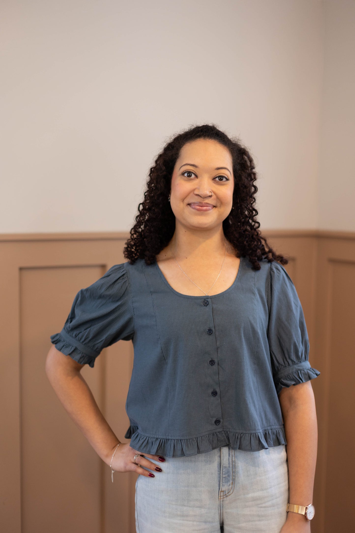 A woman with curly hair stands indoors wearing the Millie Pinstripe Top by Dogwood Cloth—a feminine blue short-sleeve blouse—paired with light jeans, one hand on her hip and a neutral expression.