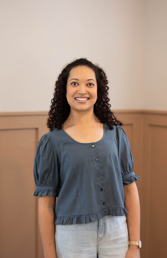A woman with curly hair smiles in a room with beige paneled walls, wearing the Dogwood Cloth Millie Pinstripe Top and light jeans.