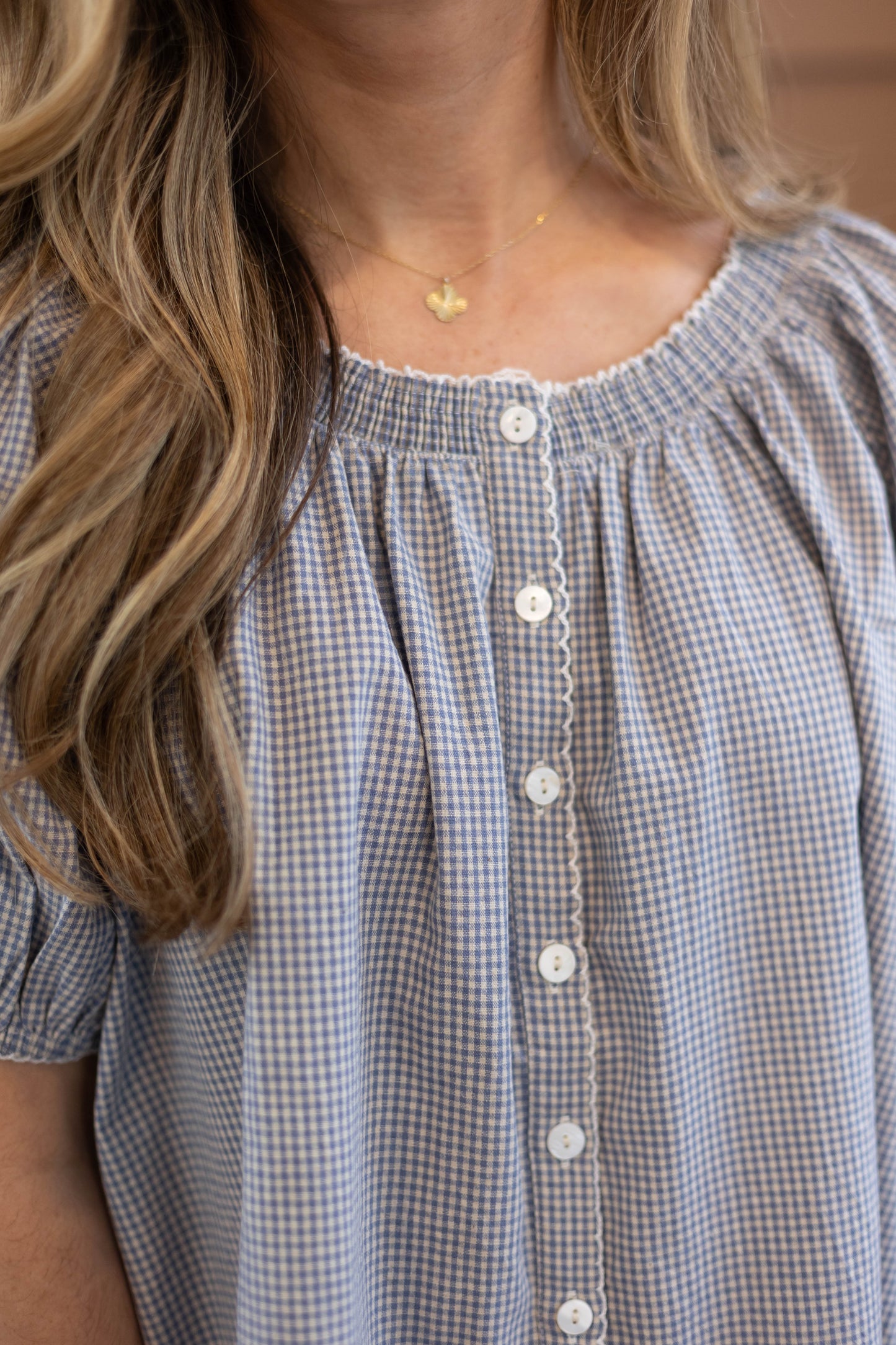 A woman wears the Dogwood Cloth Sadie Gingham Top + Scrunchie Set—a feminine blue and white check blouse with white buttons—paired with a gold pendant necklace.