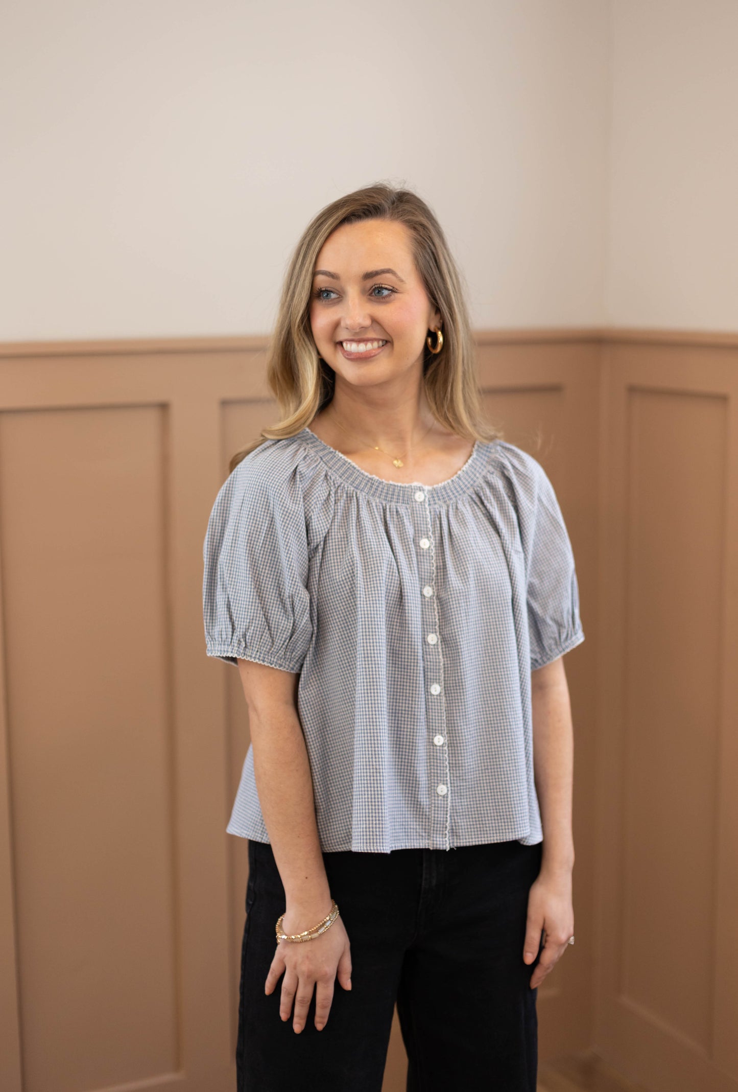 A smiling woman with long blonde hair stands indoors wearing the Dogwood Cloth Sadie Gingham Top + Scrunchie Set, a blue gingham short-sleeve button-up blouse, paired with black pants.