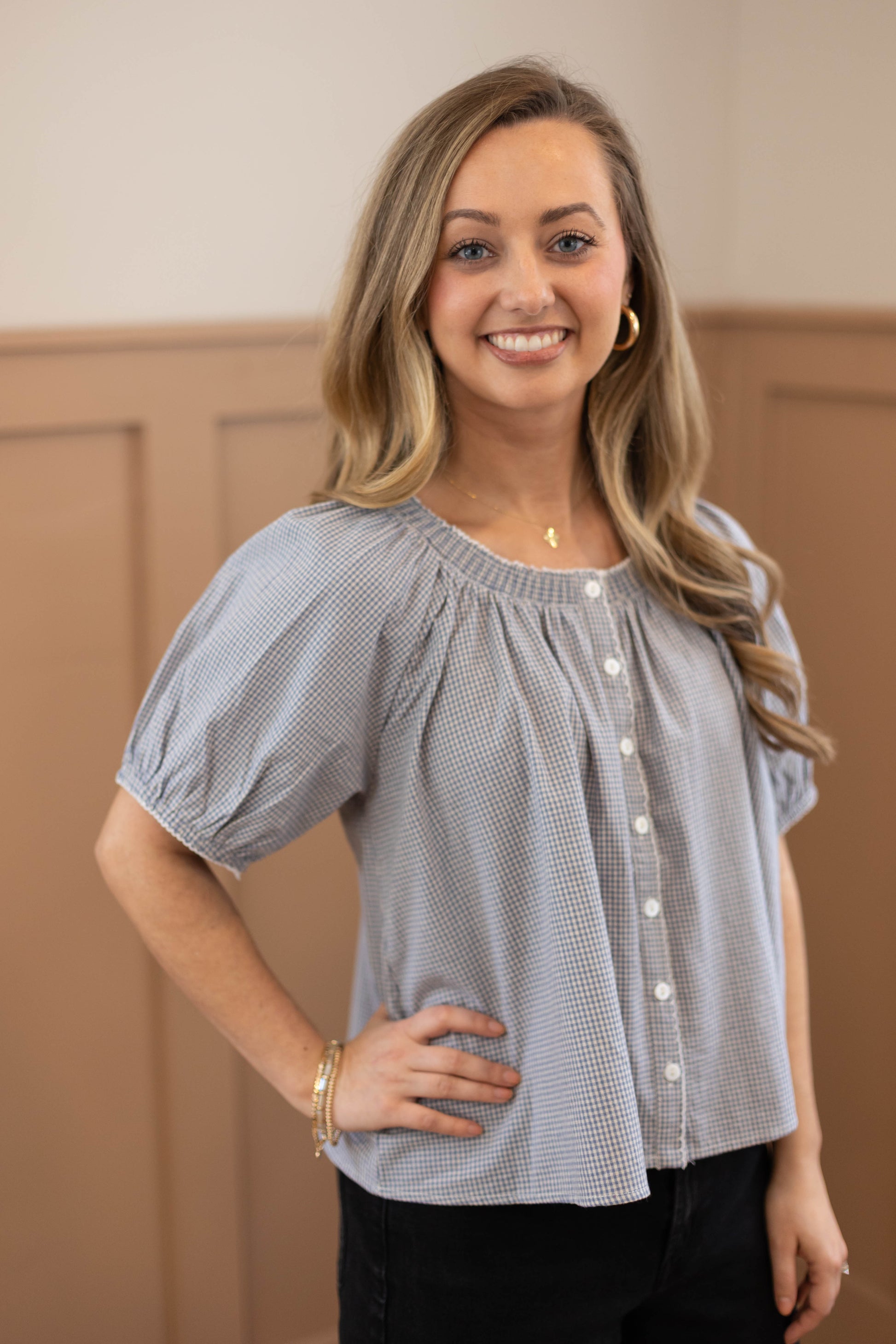 A woman with long, wavy blonde hair smiles indoors wearing the Dogwood Cloth Sadie Gingham Top + Scrunchie Set—featuring a blue gingham short-sleeve top paired with black pants, bracelets, and hoop earrings for a cheerful, stylish look.