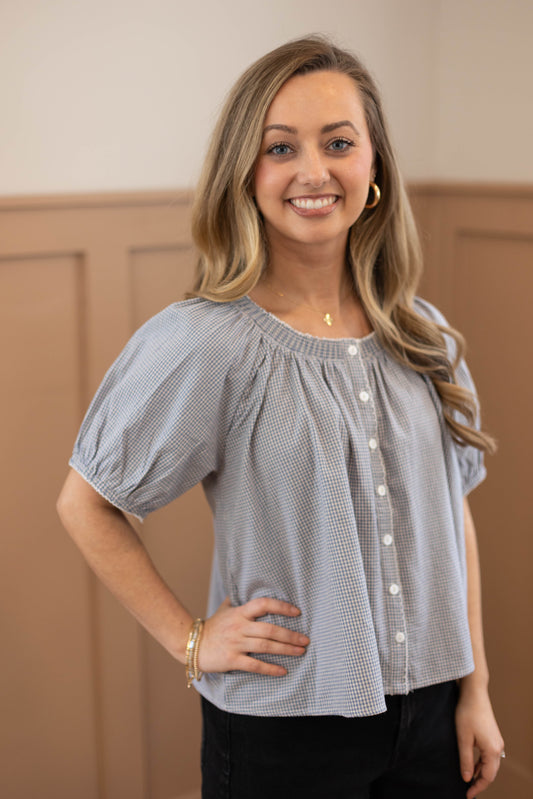 A woman with long, wavy blonde hair smiles indoors wearing the Dogwood Cloth Sadie Gingham Top + Scrunchie Set—featuring a blue gingham short-sleeve top paired with black pants, bracelets, and hoop earrings for a cheerful, stylish look.