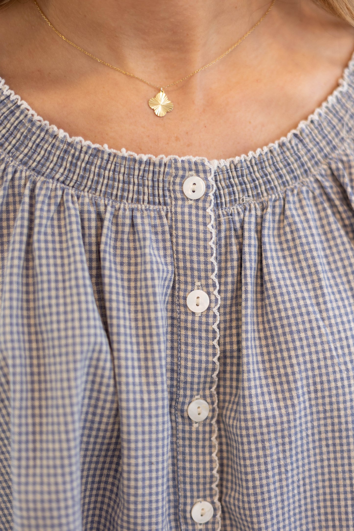 Close-up of a person wearing the Dogwood Cloth Sadie Gingham Top + Scrunchie Set, a blue and white gingham button-up blouse with scalloped trim, styled with a gold necklace featuring a small pendant.