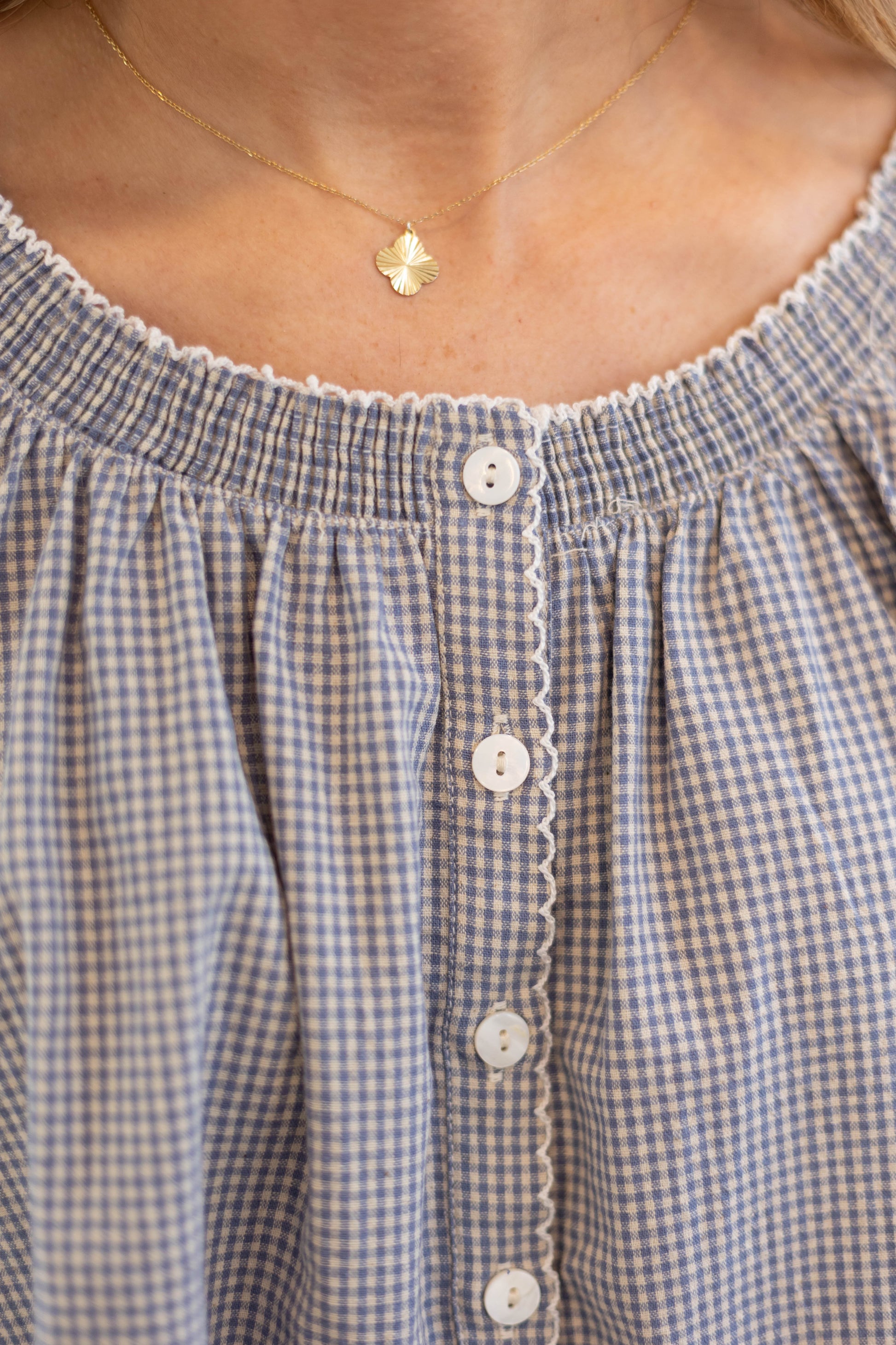 Close-up of a person wearing the Dogwood Cloth Sadie Gingham Top + Scrunchie Set, a blue and white gingham button-up blouse with scalloped trim, styled with a gold necklace featuring a small pendant.