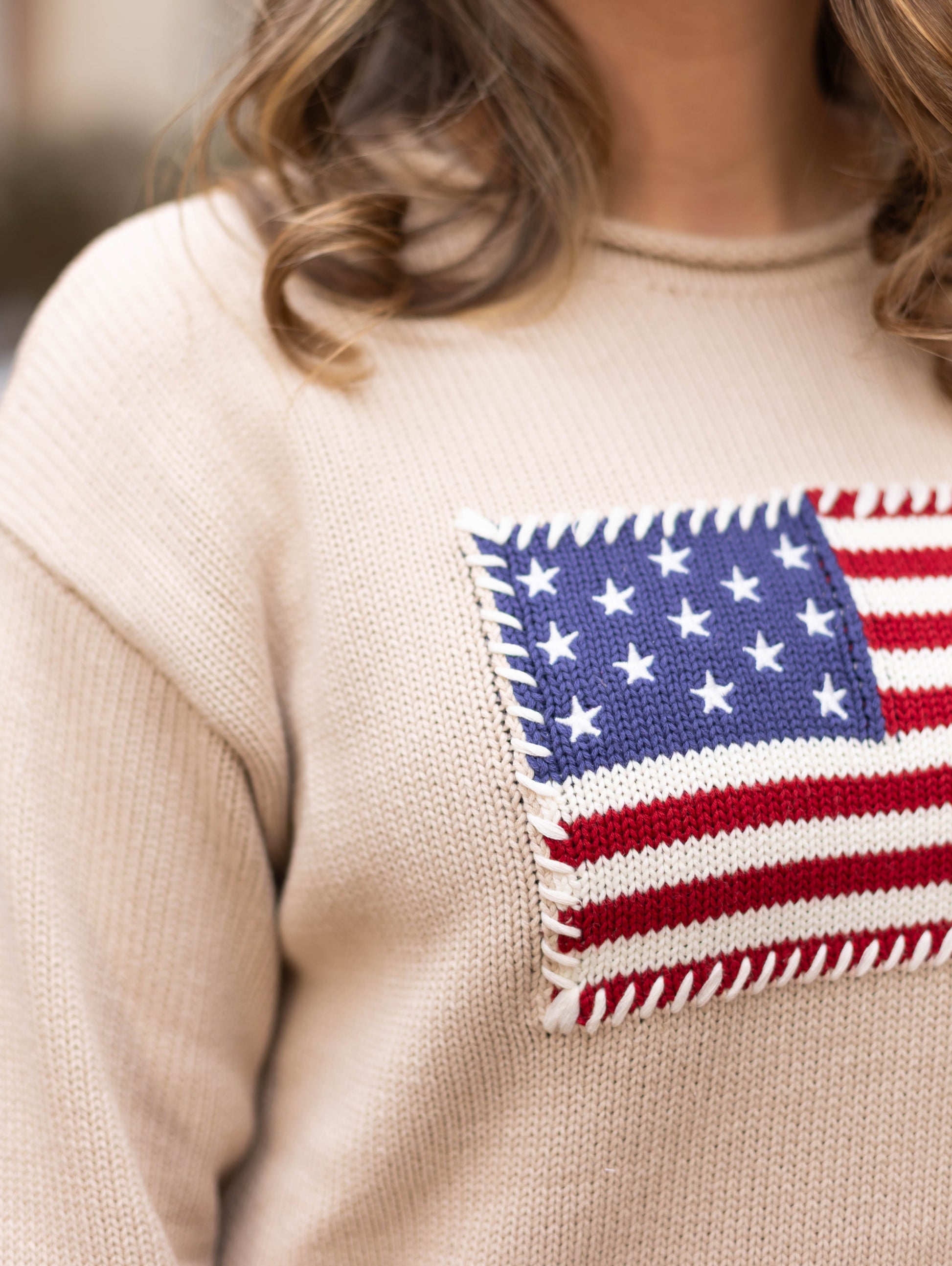 A person wearing the Dogwood Cloth Heirloom Knit Sweater—made from 100% cotton with a knitted American flag patch on the front. Only their upper body is shown.
