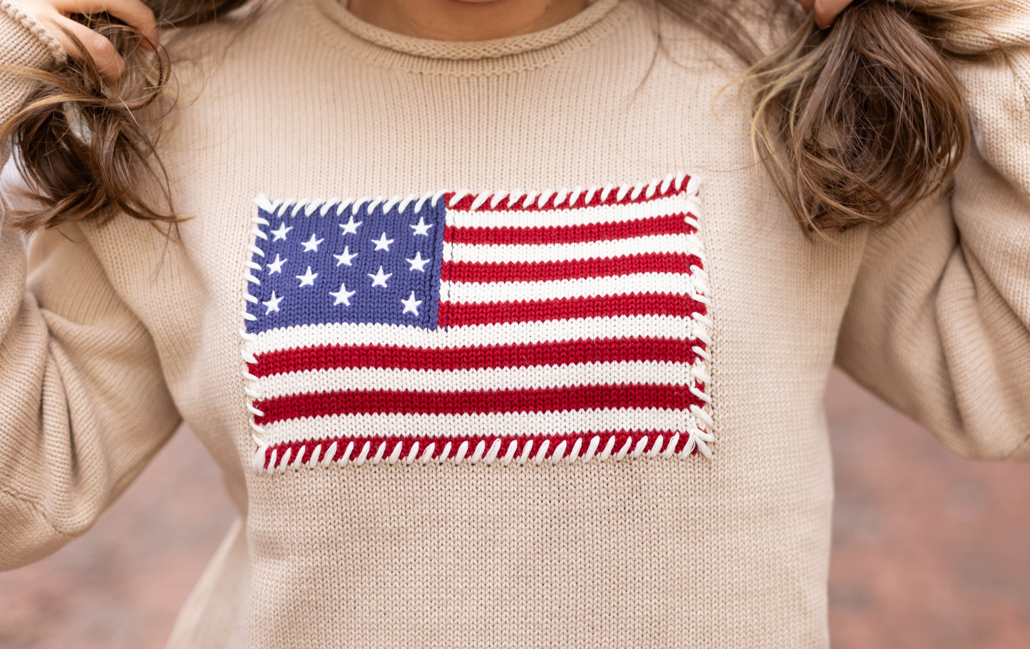 A person wearing the Dogwood Cloth Heirloom Knit Sweater, featuring an embroidered American flag patch on the front, holds sections of their hair.