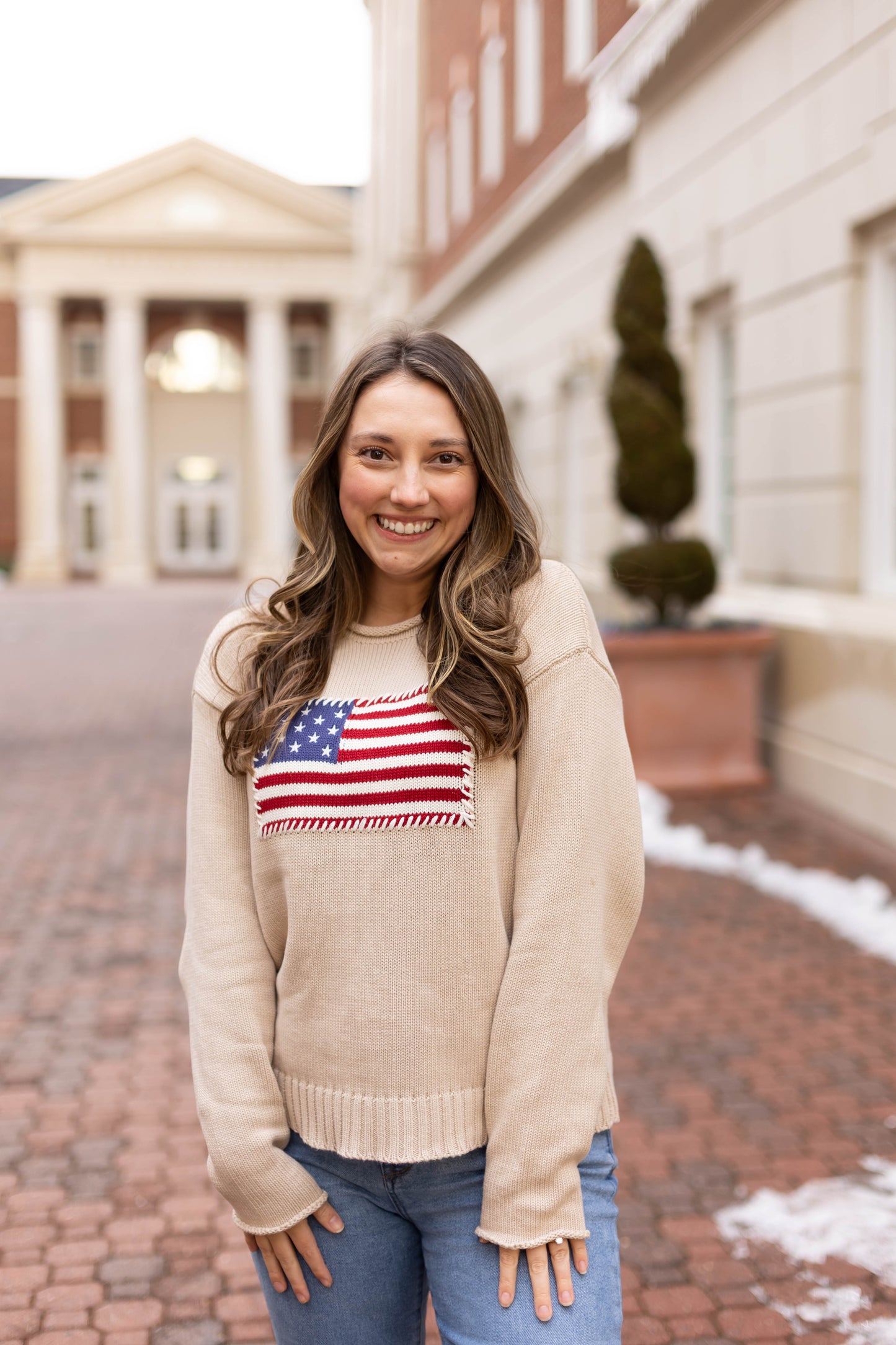 A young woman smiles at the camera outdoors, wearing Dogwood Cloth’s Heirloom Knit Sweater—an American flag design crafted from 100% cotton—paired with blue jeans.
