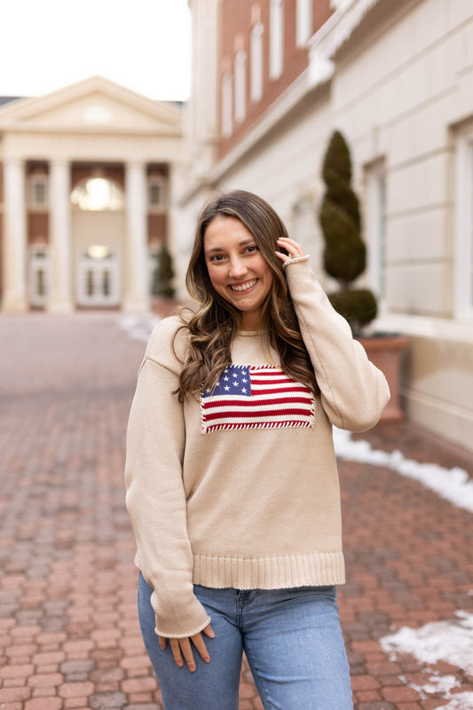 A woman smiles outside on a brick walkway, wearing Dogwood Cloth’s Heirloom Knit Sweater—an American flag design made from 100% cotton—with buildings in the background.