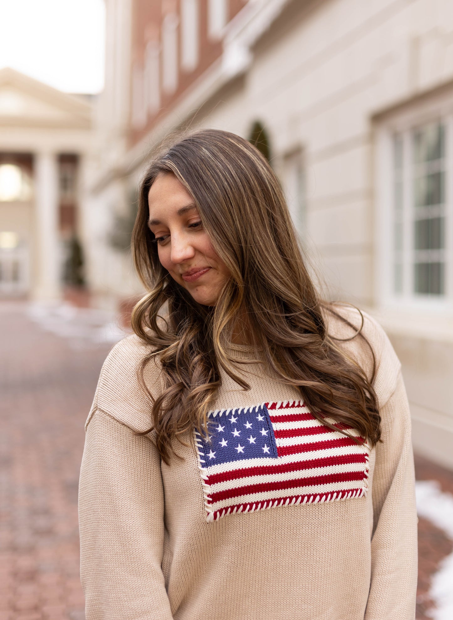 A woman with long brown hair wears the Dogwood Cloth Heirloom Knit Sweater with an American flag patch, standing on a brick pathway next to a light-colored building.