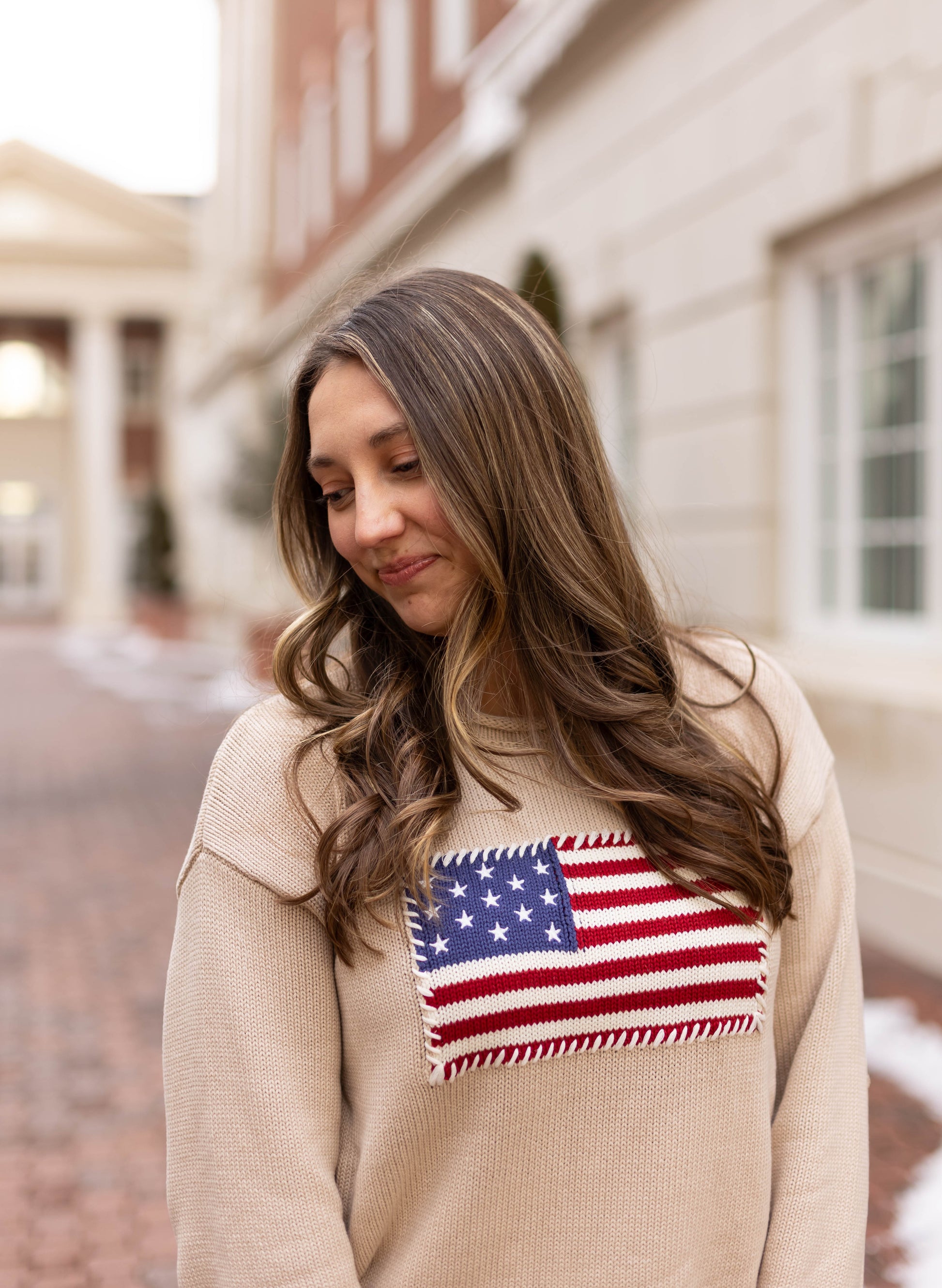 A woman with long brown hair wears the Dogwood Cloth Heirloom Knit Sweater with an American flag patch, standing on a brick pathway next to a light-colored building.