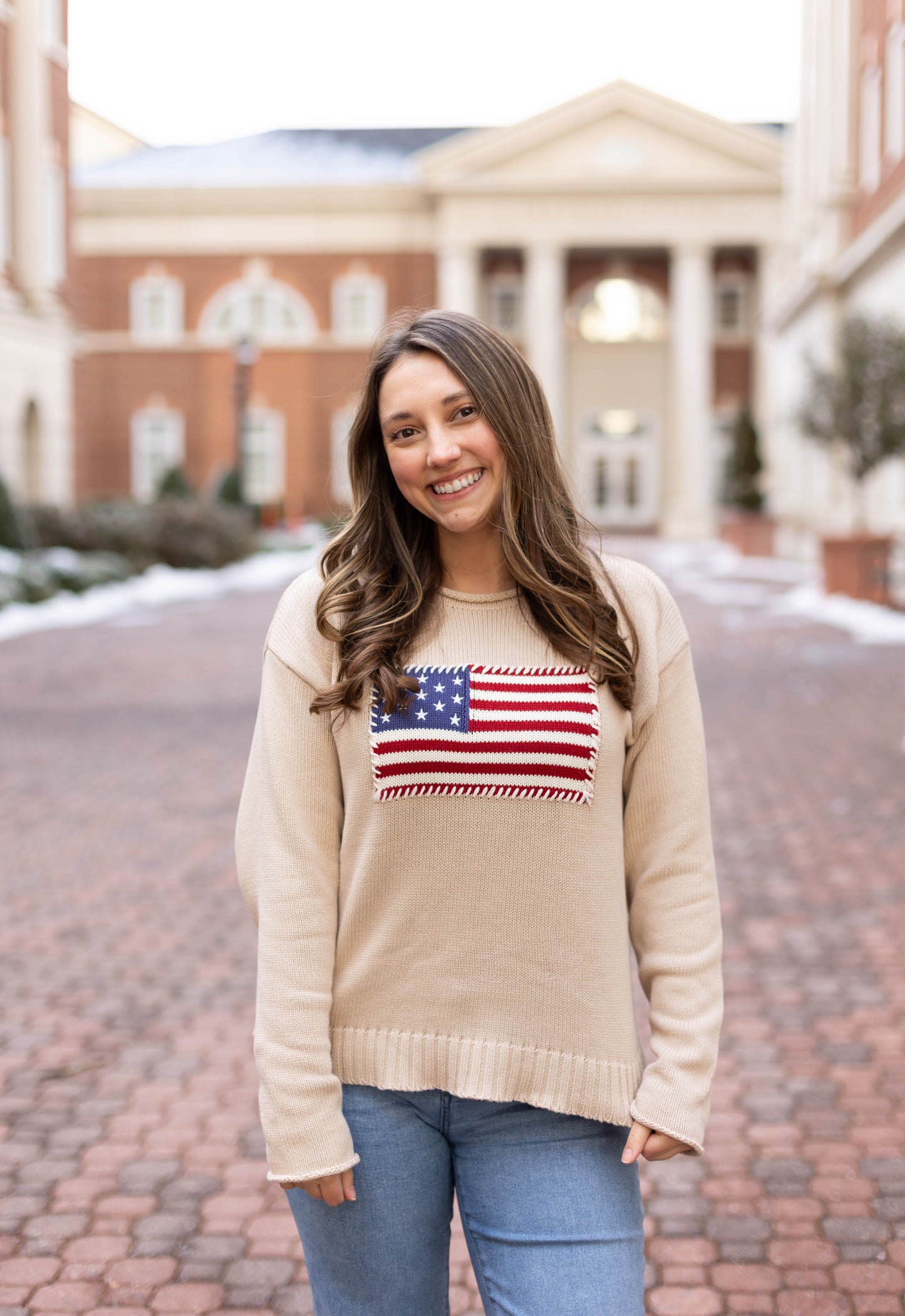 A woman smiles on a brick walkway in front of a columned building, wearing blue jeans and the Dogwood Cloth Heirloom Knit Sweater featuring an American flag, made from 100% cotton.