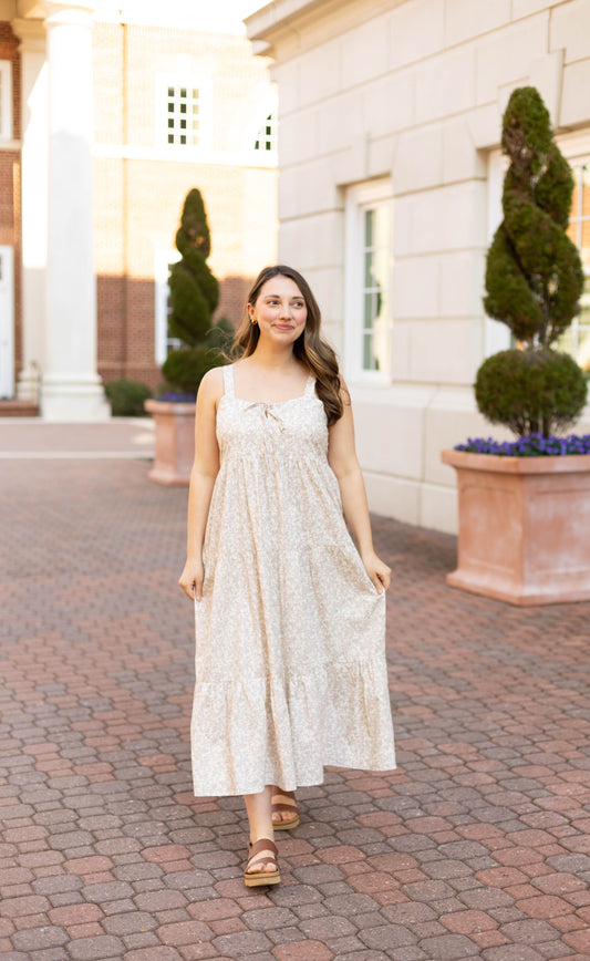 A woman in the Avery Floral Maxi by Dogwood Cloth strolls along a brick pathway lined with potted topiary trees and flowers between buildings.