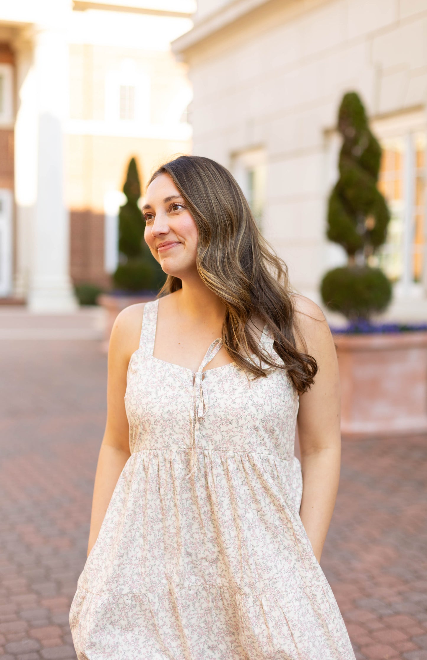 A woman with long brown hair smiles slightly outdoors on a brick walkway, wearing the Avery Floral Maxi dress by Dogwood Cloth.