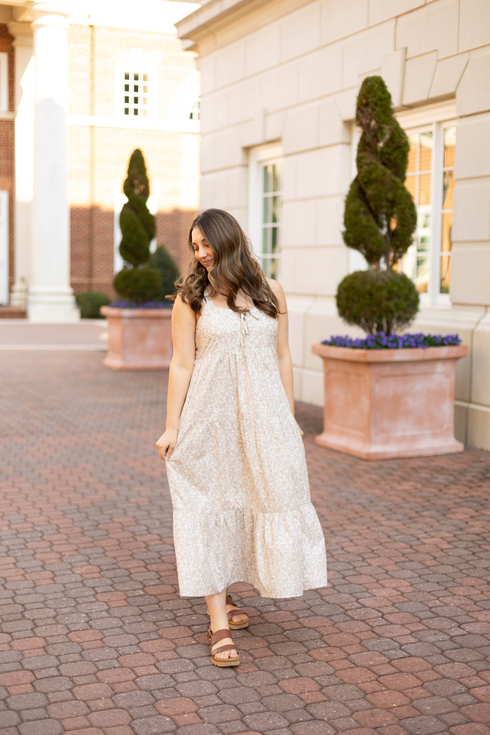 Wearing the Avery Floral Maxi by Dogwood Cloth, a woman strolls along a brick path near potted plants and large windows, showing off the dress’s relaxed fit and effortlessly chic style.
