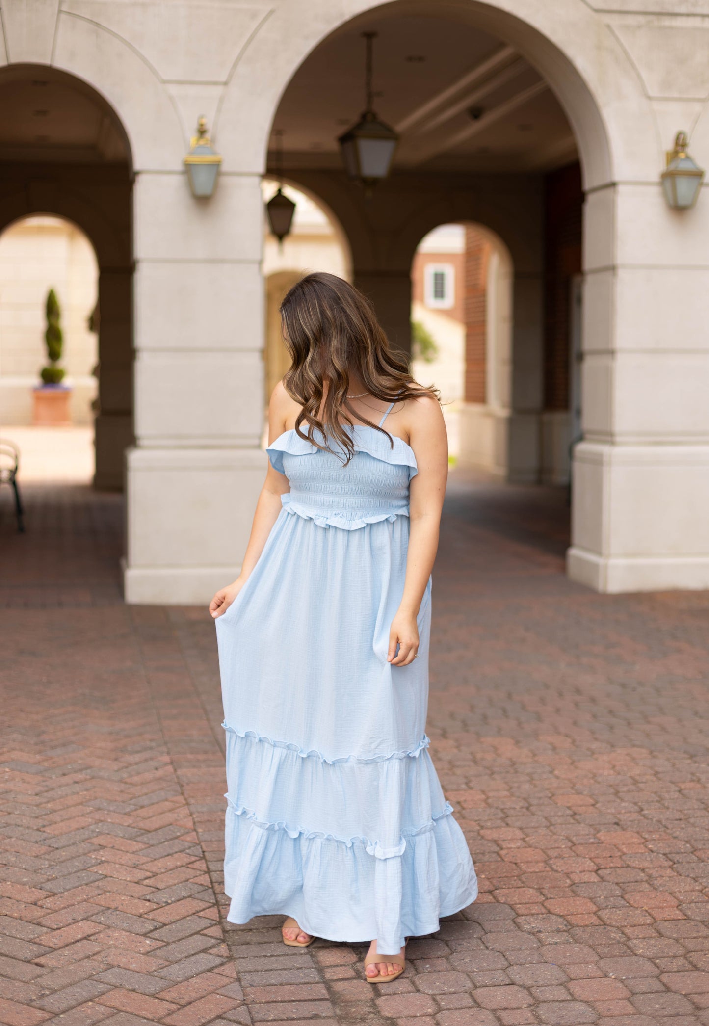 A woman in the Dogwood Cloth Seabrook Tiered Maxi stands on a brick path under an arched walkway, looking down and holding her sky blue dress.