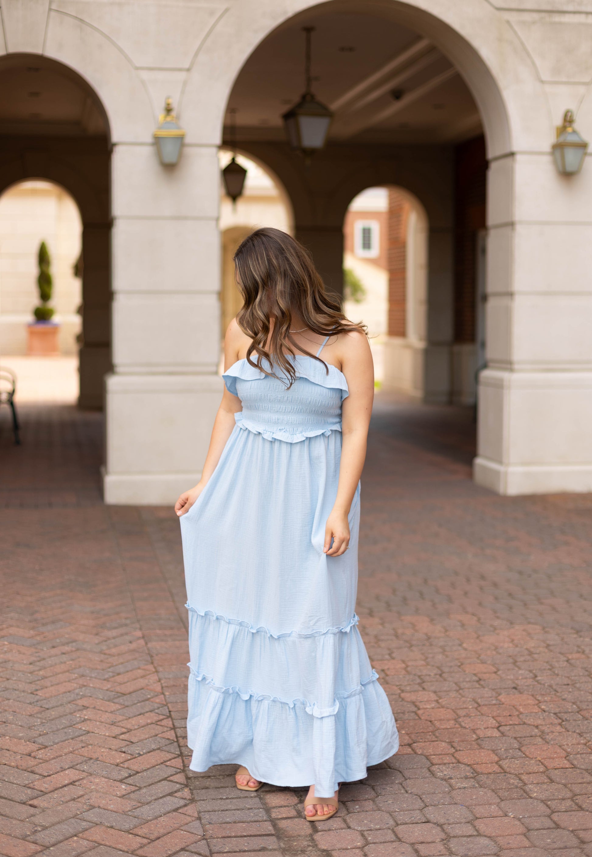 A woman in the Dogwood Cloth Seabrook Tiered Maxi stands on a brick path under an arched walkway, looking down and holding her sky blue dress.