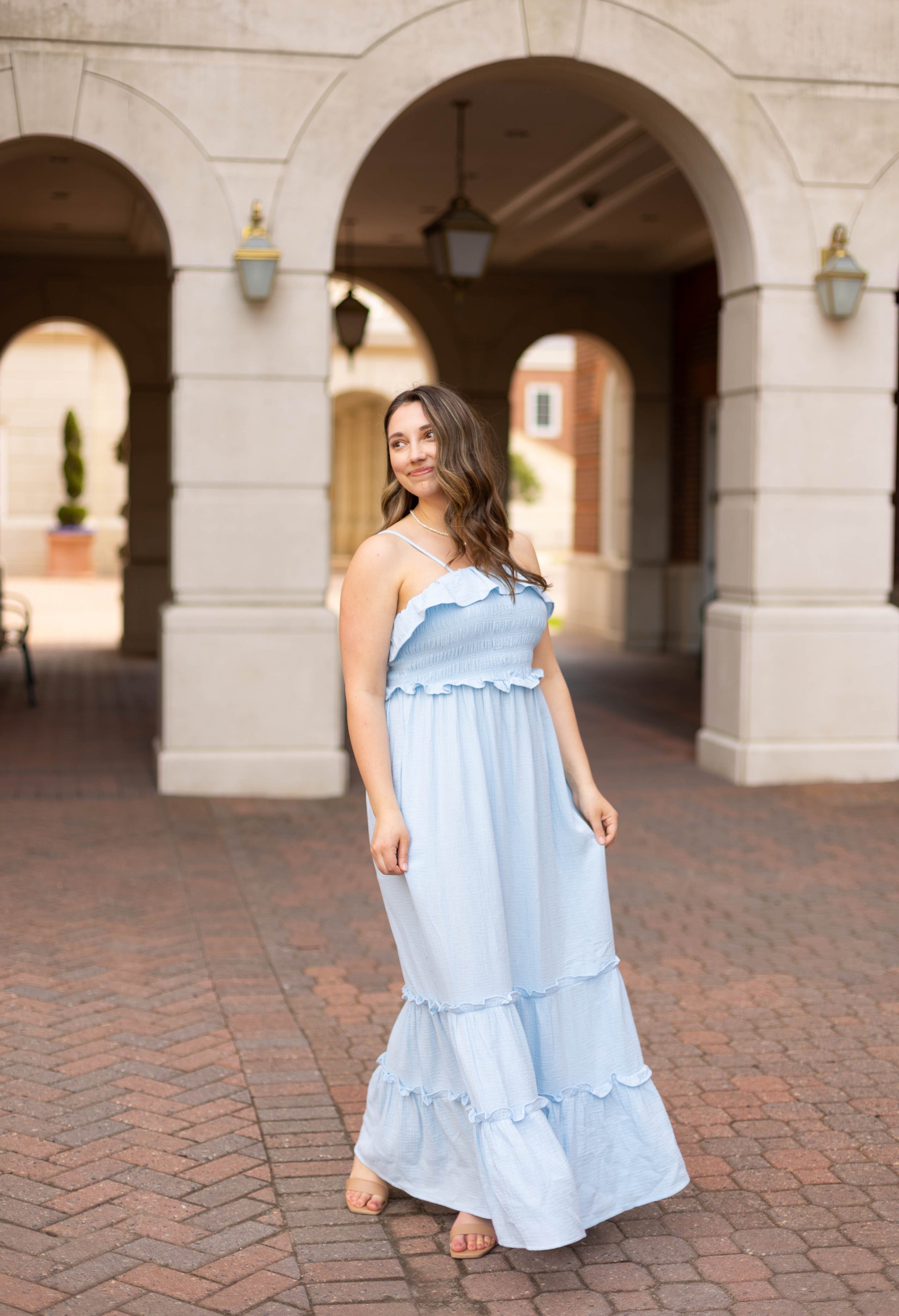 A woman in the Dogwood Cloth Seabrook Tiered Maxi, a light blue dress, stands on a brick walkway by an arched building, gazing off to the side.