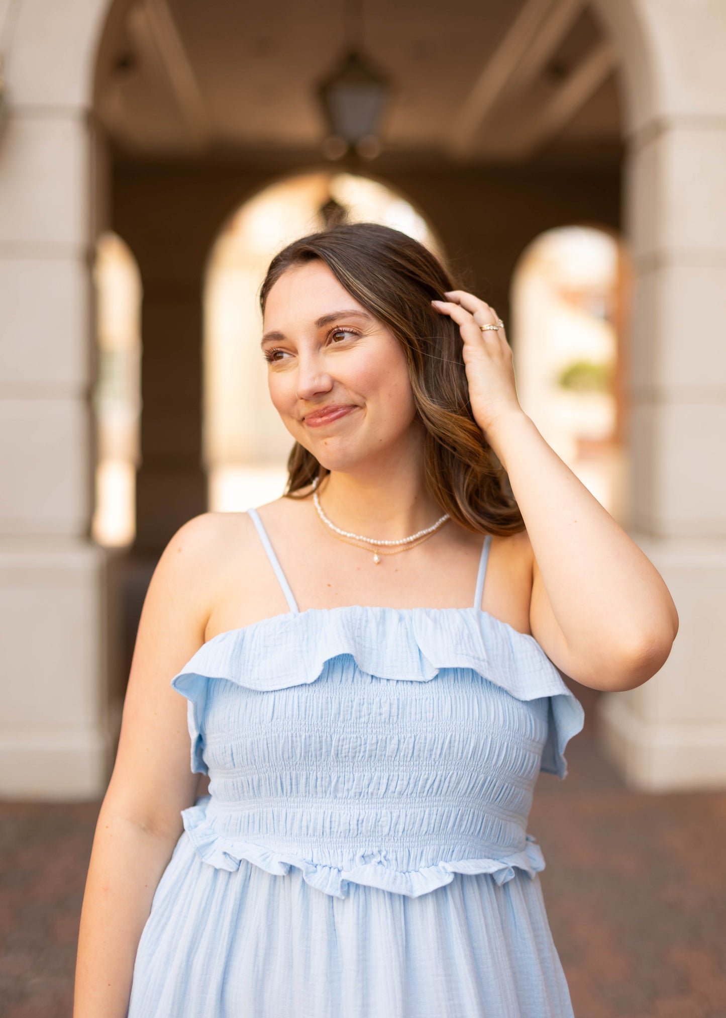 A woman wearing the Dogwood Cloth Seabrook Tiered Maxi in sky blue stands outside under an archway, smiling and touching her hair.