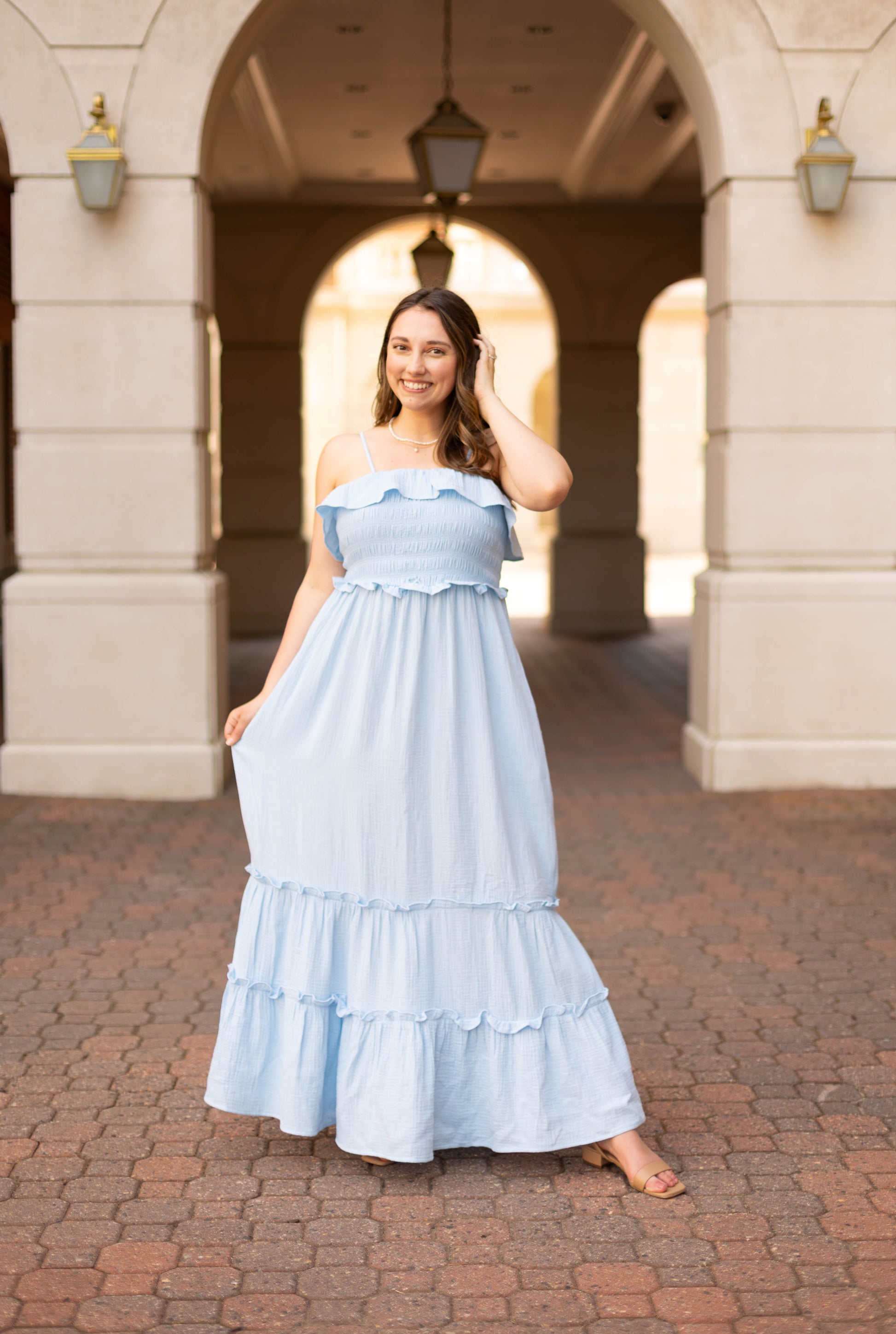 A woman stands outdoors on a brick pathway wearing the Dogwood Cloth Seabrook Tiered Maxi in sky blue, holding her dress with one hand and touching her hair with the other.