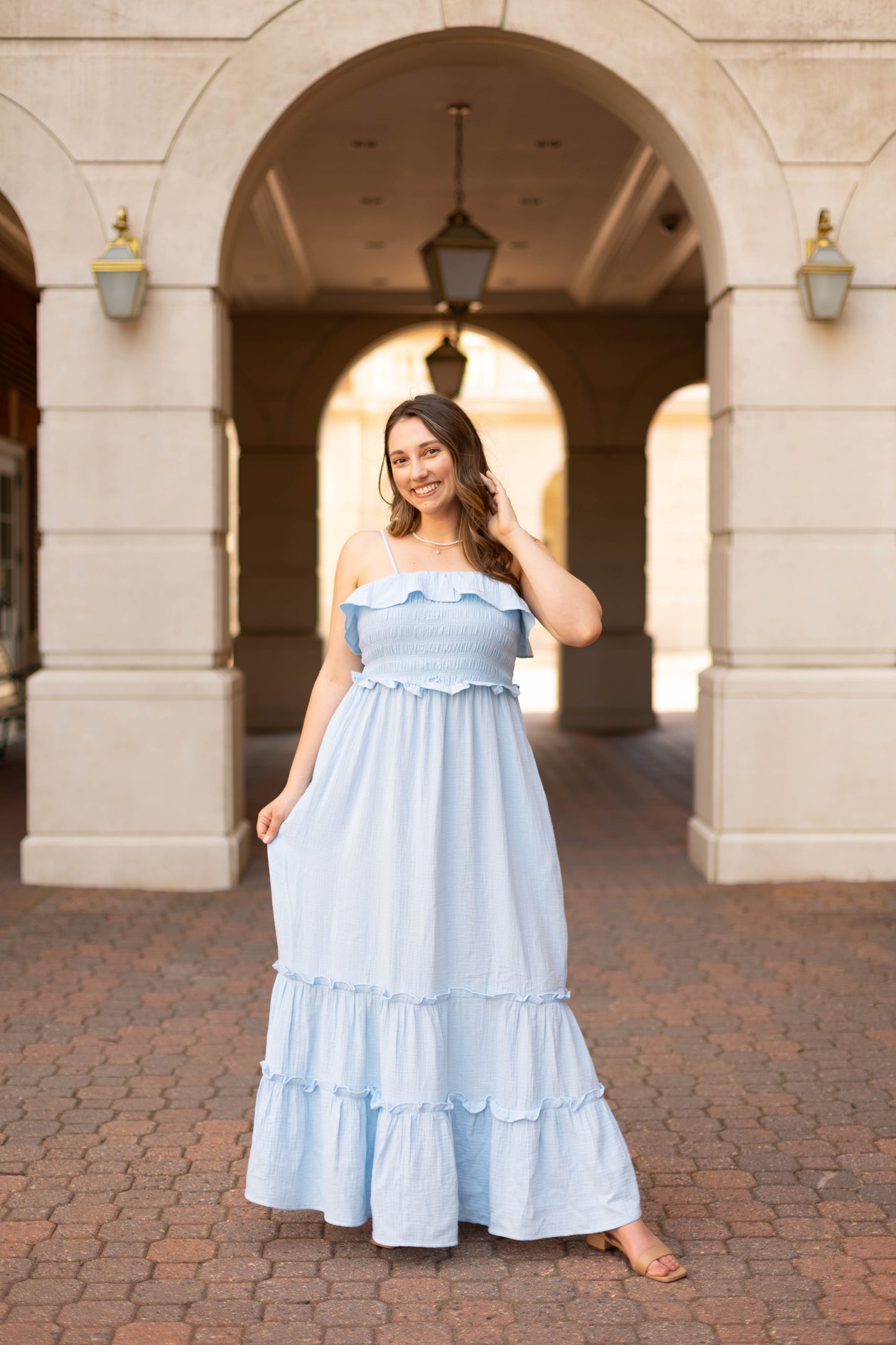 A woman smiles outdoors under stone arches, touching her hair while wearing the Dogwood Cloth Seabrook Tiered Maxi in sky blue with an off-shoulder design.
