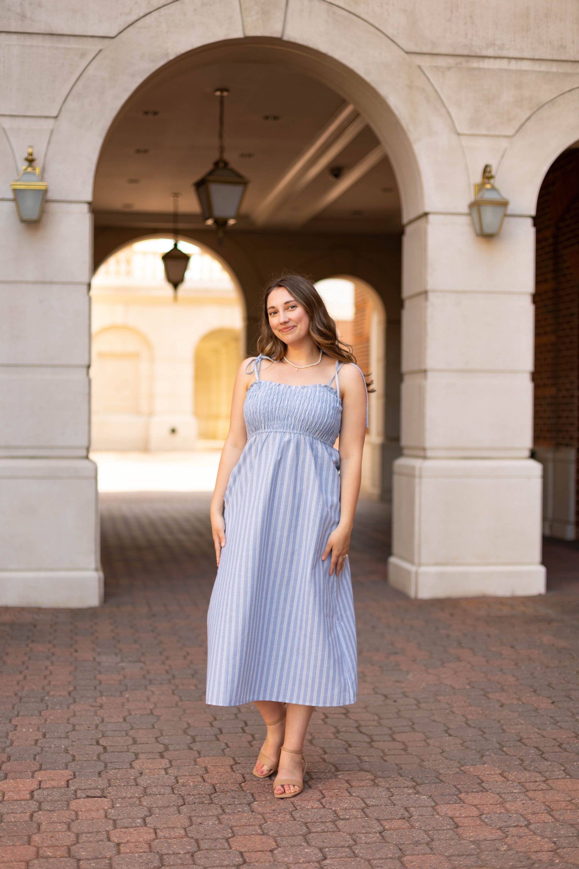 A woman smiles at the camera under a stone archway with lanterns, modeling the Amelia Striped Midi by Dogwood Cloth—a chic blue and white cotton dress with standout style details.