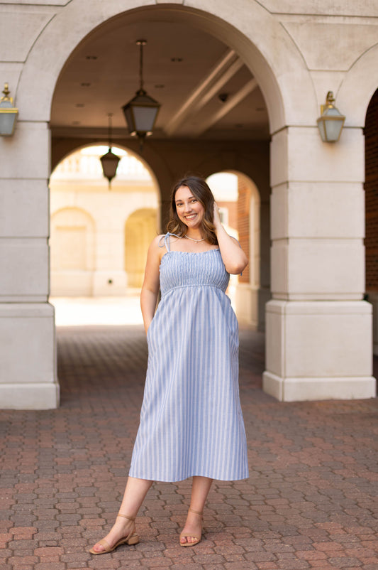 A woman smiles at the camera under an archway, wearing the Dogwood Cloth Amelia Striped Midi, a blue and white 100% cotton sundress with fit details, paired with tan sandals and her hand in her hair.