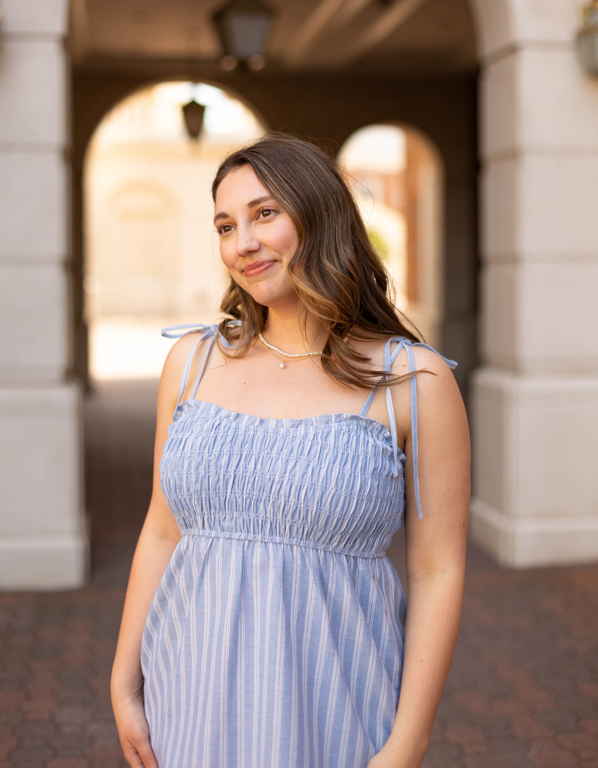 A woman stands outdoors in front of an arched building, smiling slightly while wearing the Amelia Striped Midi by Dogwood Cloth—a light blue, sleeveless cotton dress with stripes.